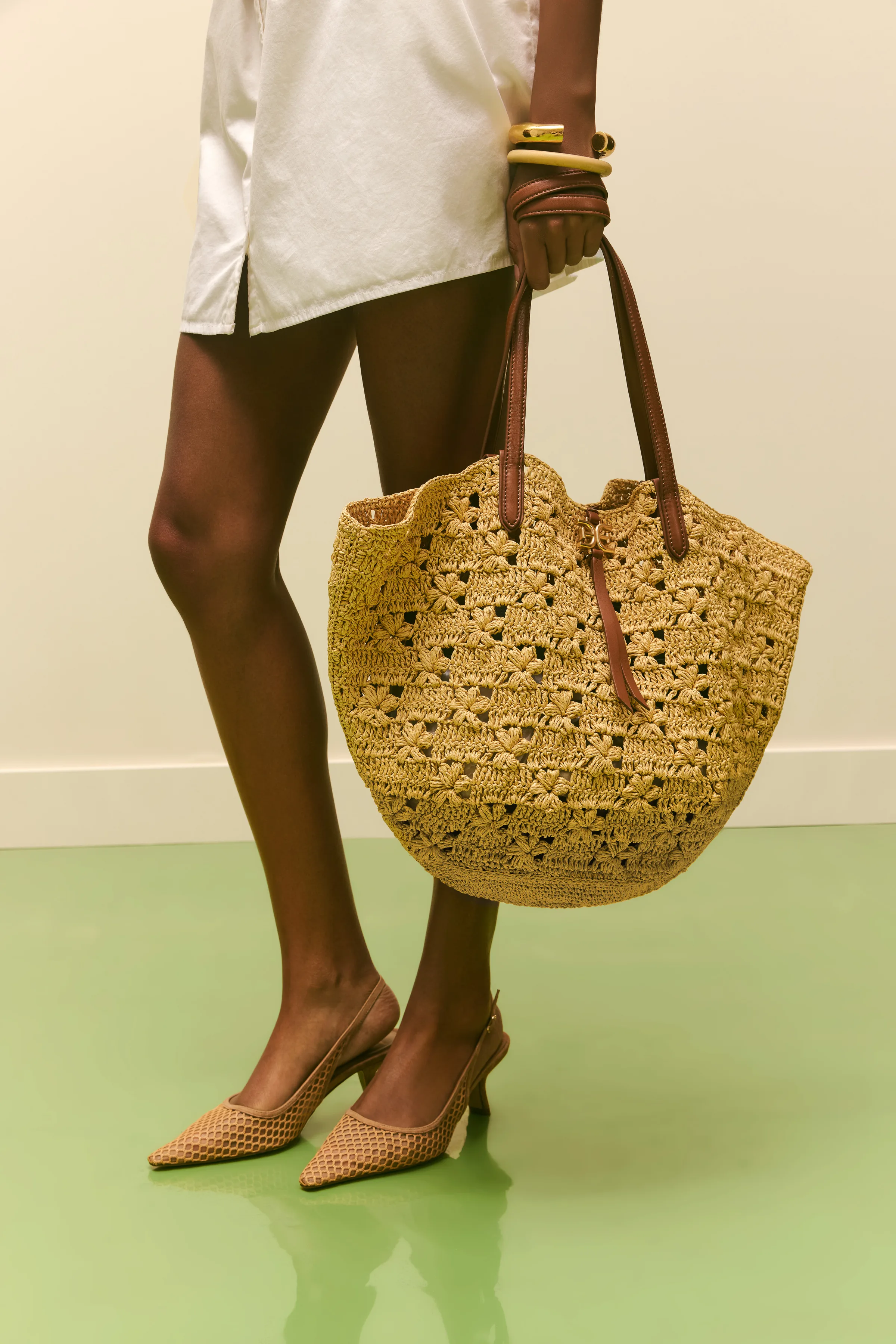 Model holding a woven straw crochet tote, wearing tan mesh slingback heels on a glossy green floor, for Sam Edelman.