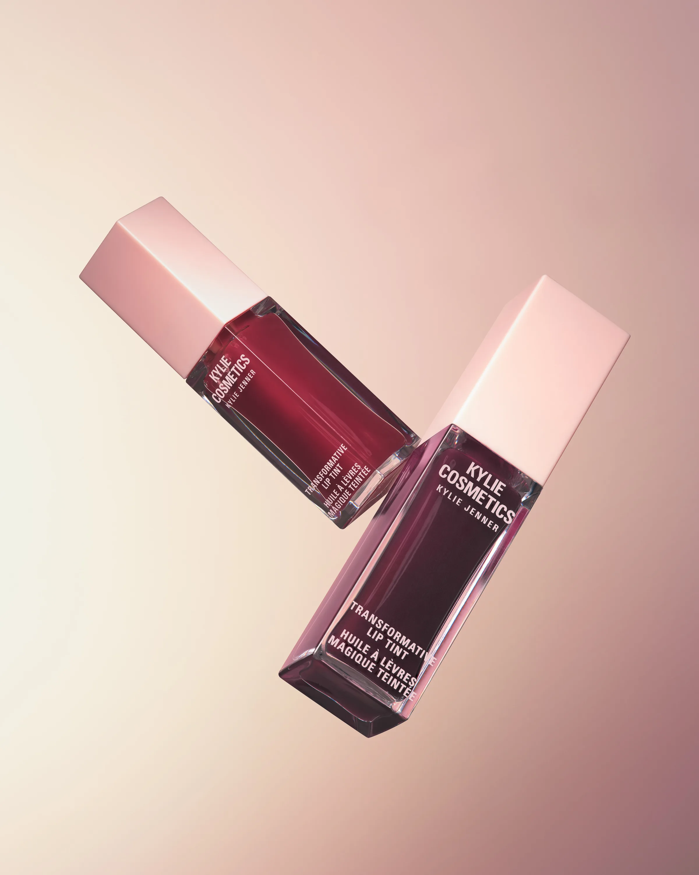 Two Transformative Lip Tint bottles in red and plum with pink caps floating on a peach gradient, for Kylie Cosmetics.
