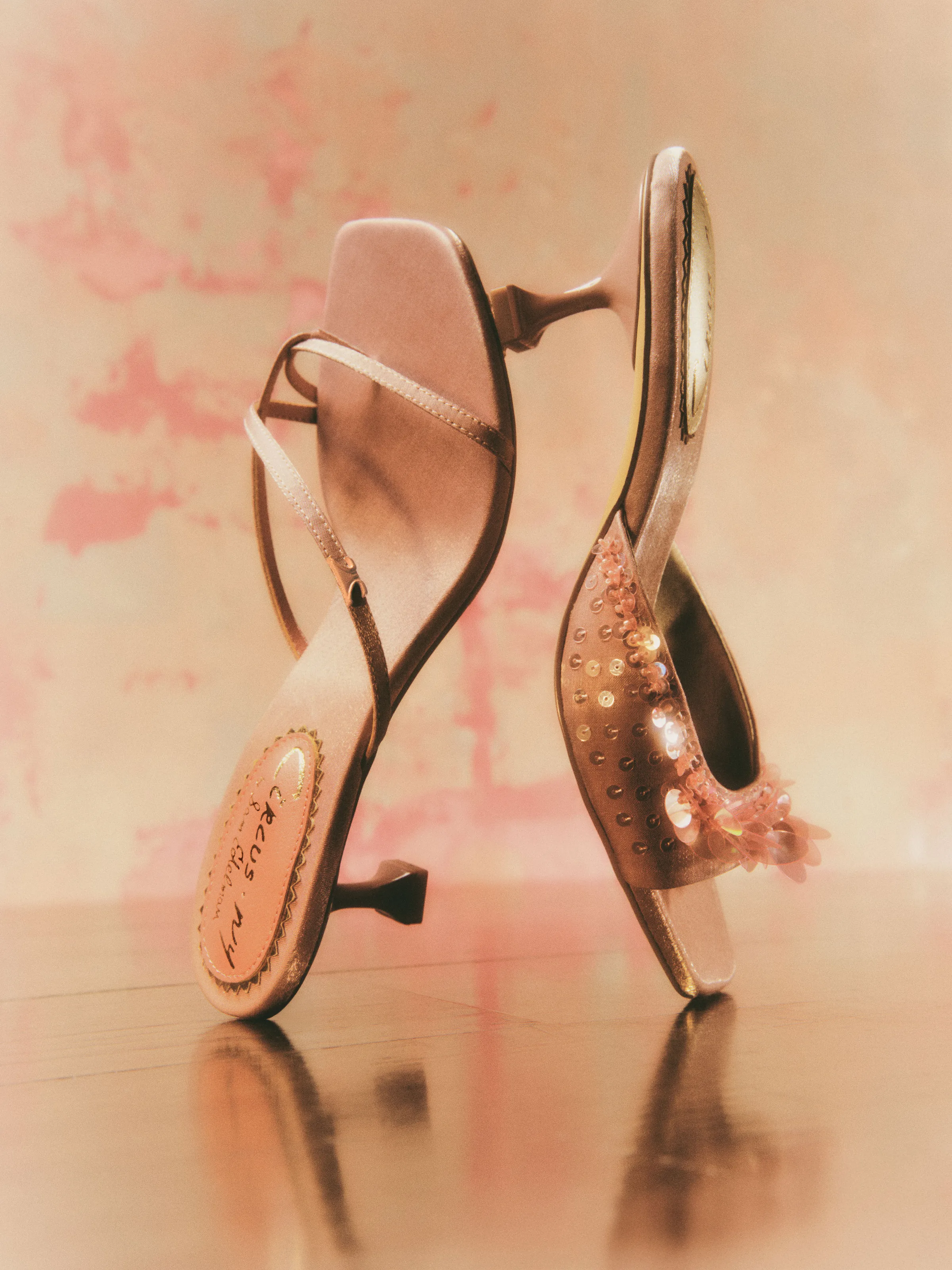 Blush satin kitten heel sandals, one strappy and one sequin-embellished, on a glossy floor against a pink wall, for Circus NY.