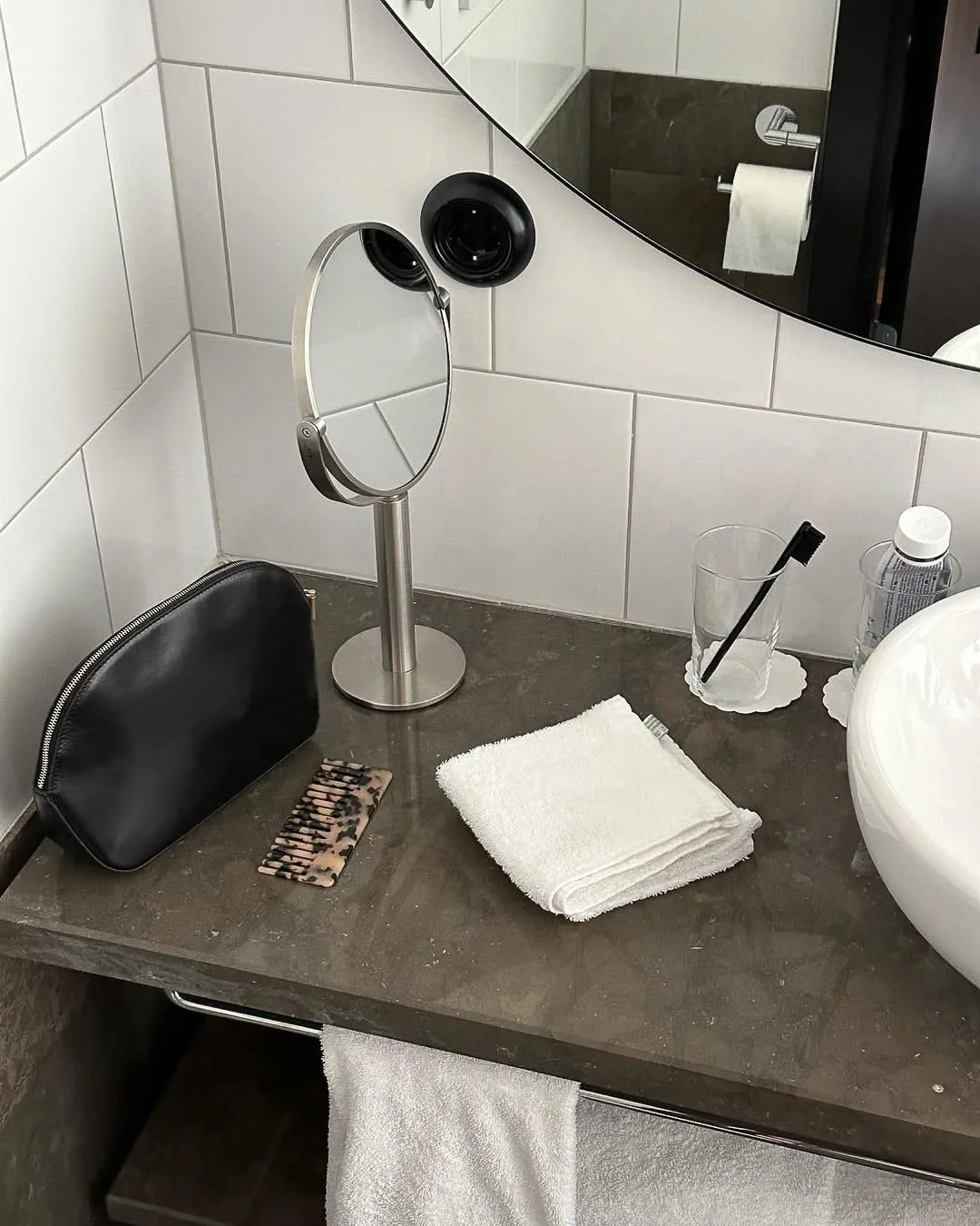 Bathroom vanity with a black leather toiletry bag, tortoiseshell comb, folded white towel and round chrome mirror.