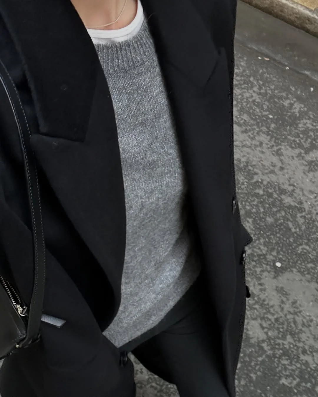 Phone-style outfit shot looking down at a black wool coat over a grey knit sweater, with a black leather bag on the street.