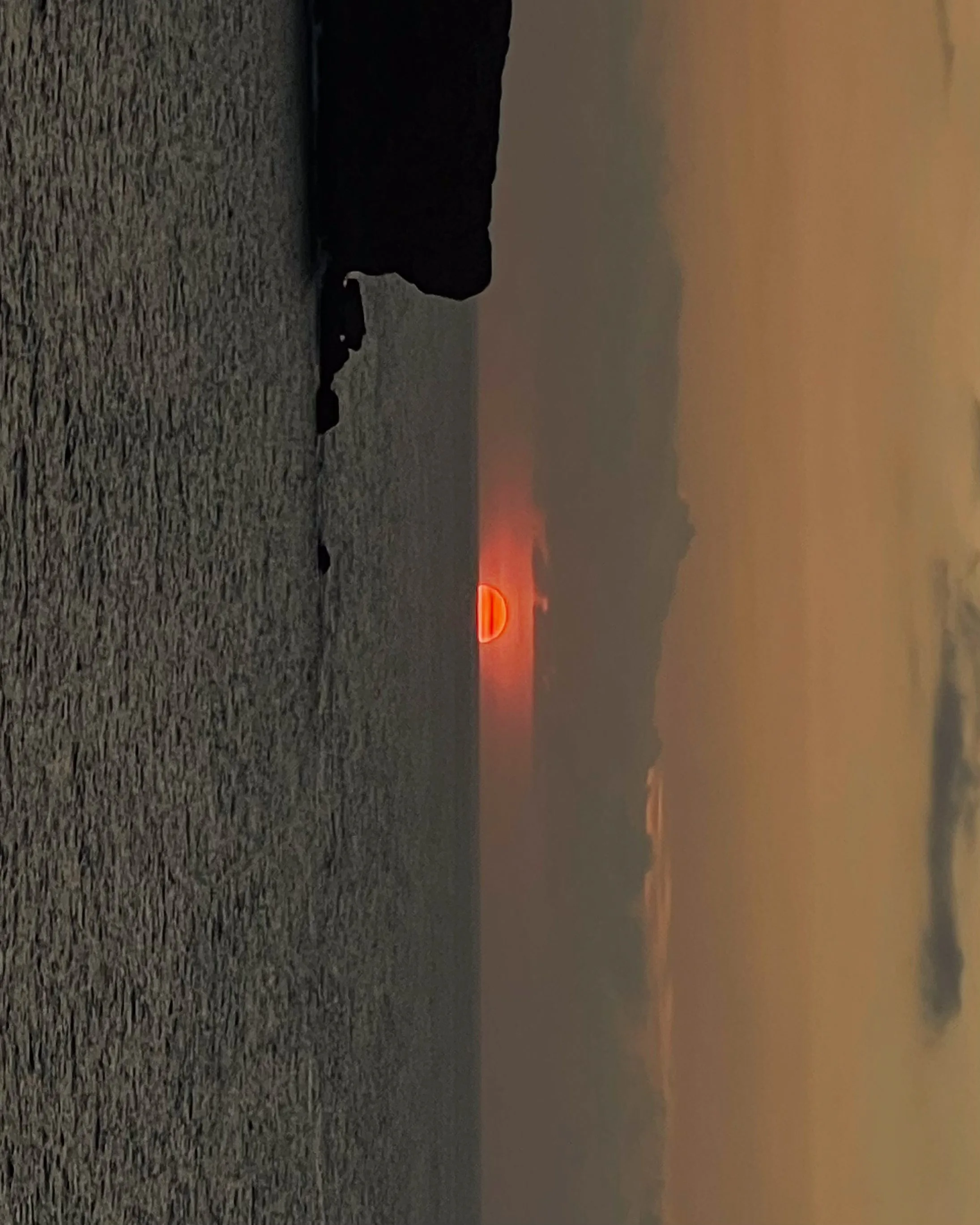 Red sun setting over a hazy sea beside a dark rocky cliff, shown rotated sideways in a phone-style shot.