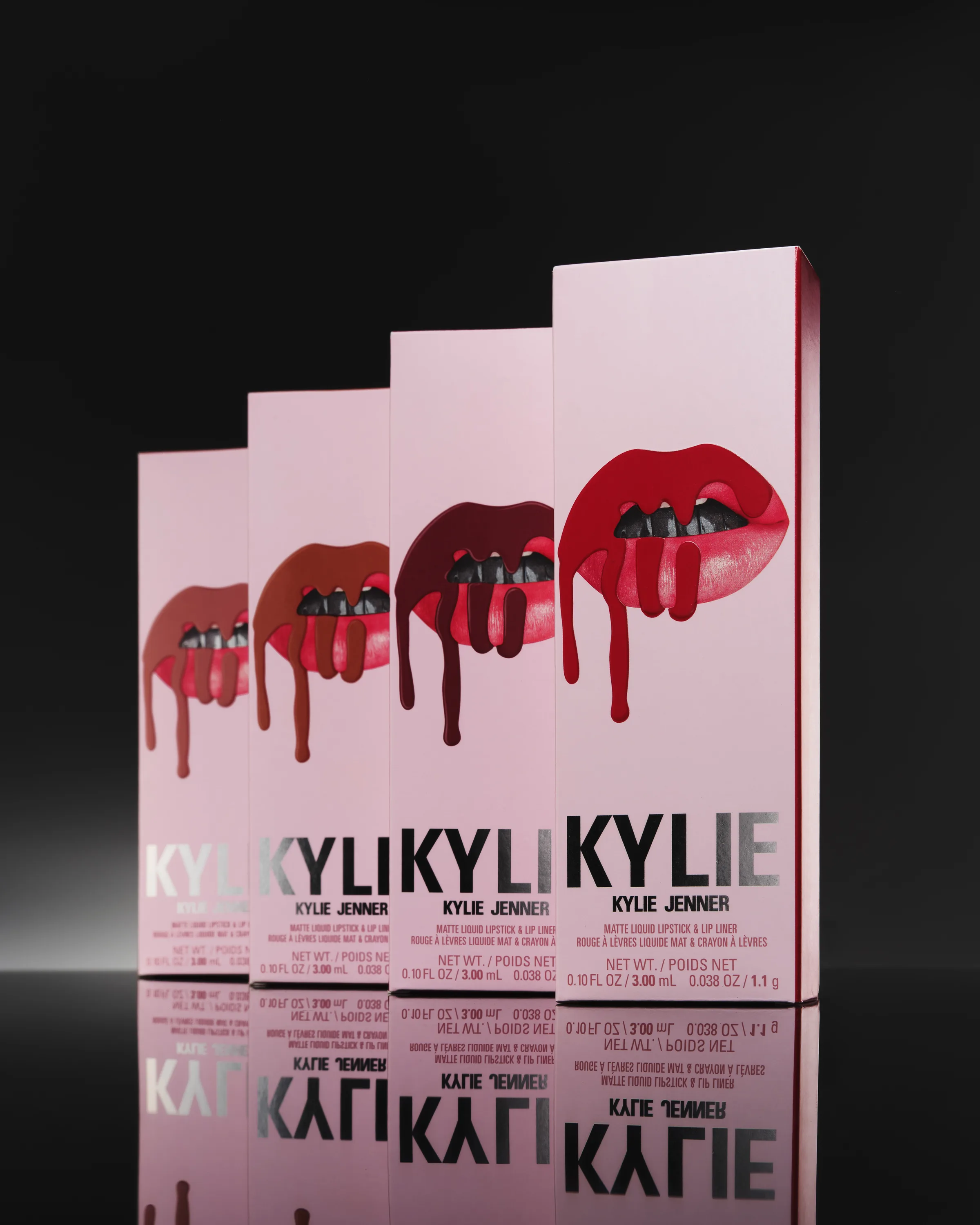 Four pink Kylie lip kit boxes with dripping lip graphics in a row on a reflective black surface, for Kylie Cosmetics.