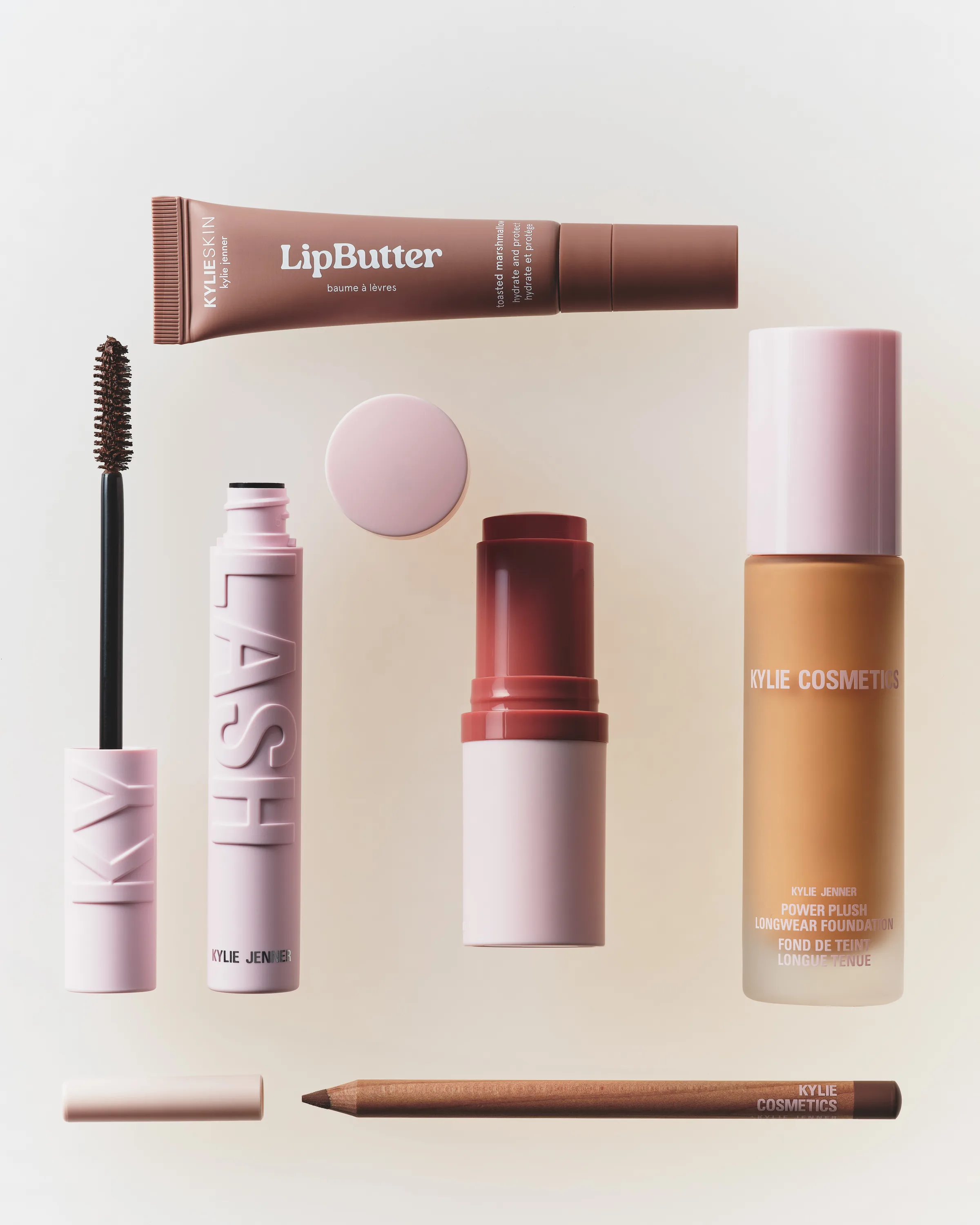 Flat lay of pink Kylie mascara, lip butter tube, blush stick, foundation bottle and lip liner on cream, for Kylie Cosmetics.
