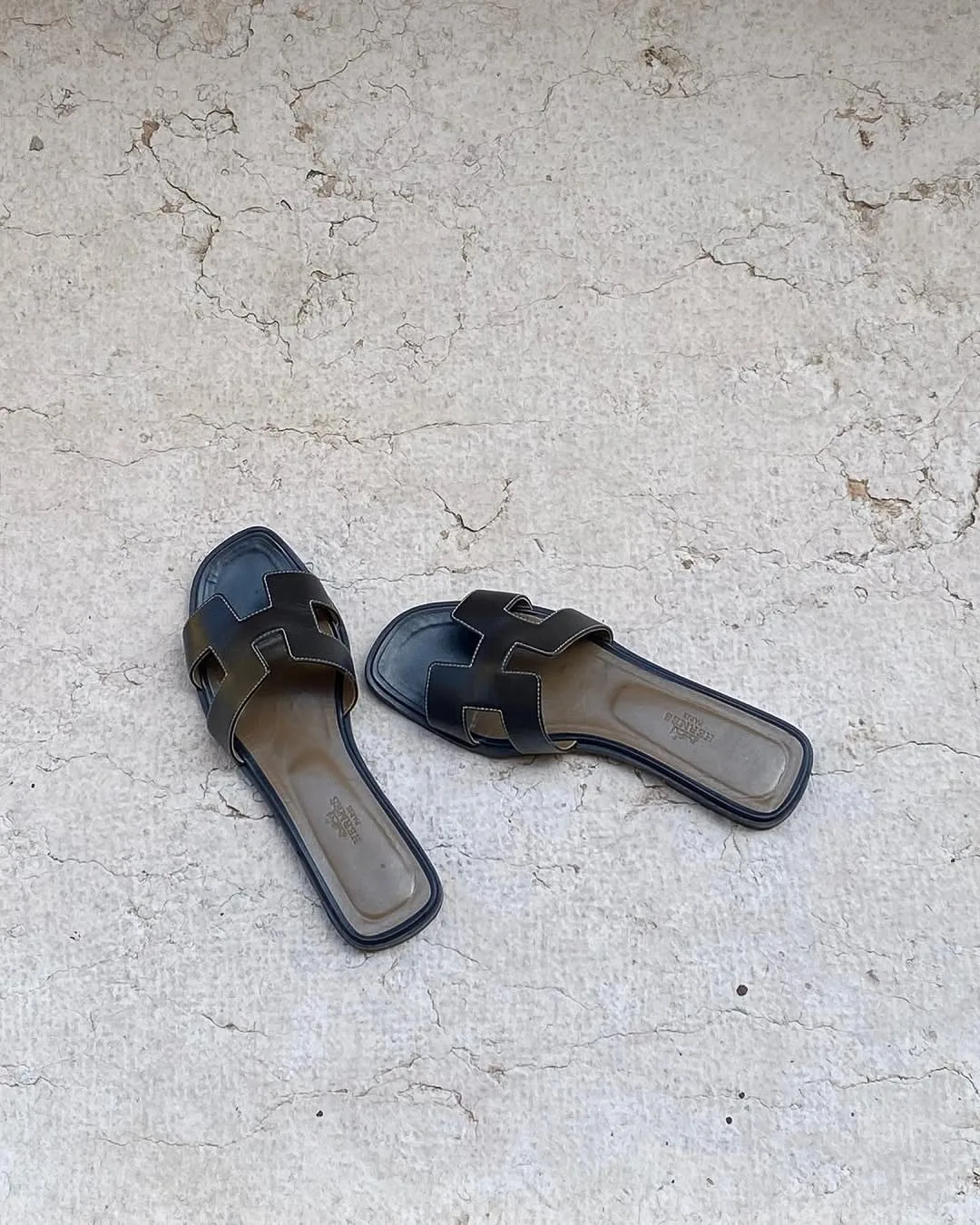 Worn black leather H-strap slide sandals on a cracked white plaster floor, shot from above in phone-camera style.