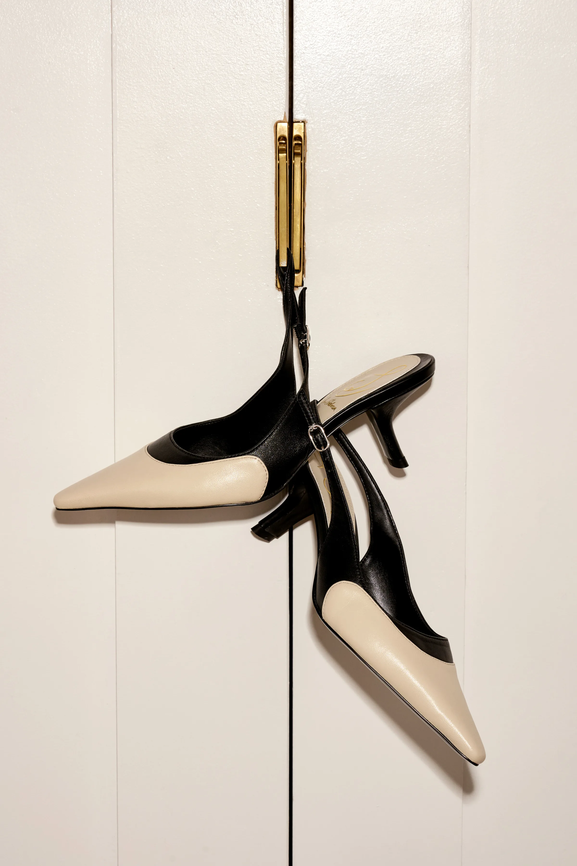Cream and black two-tone slingback kitten heels hanging from a brass latch on white cabinet doors, for Sam Edelman.