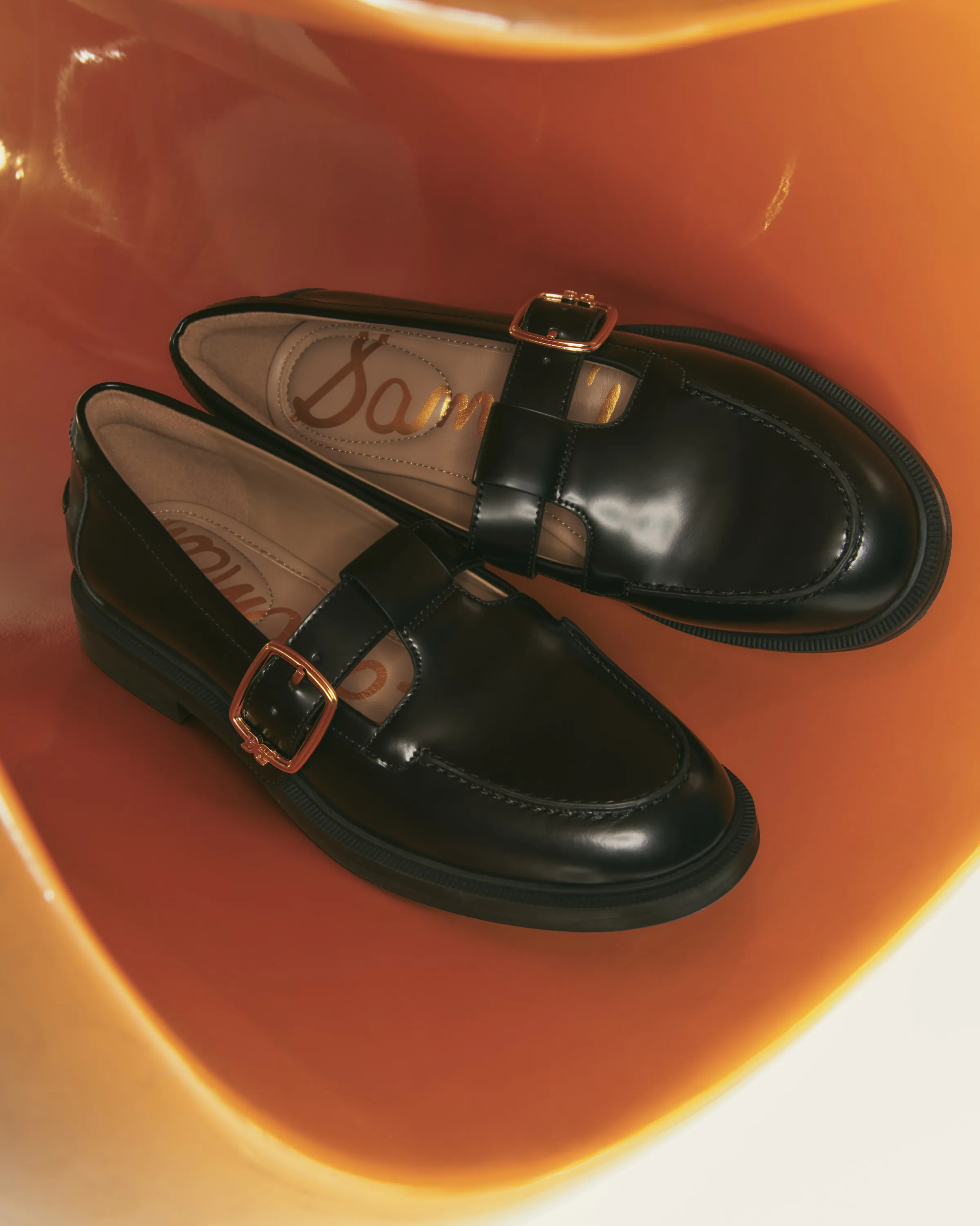 Black patent leather loafers with gold buckle straps resting in a glossy orange sculptural bowl, for Sam Edelman.