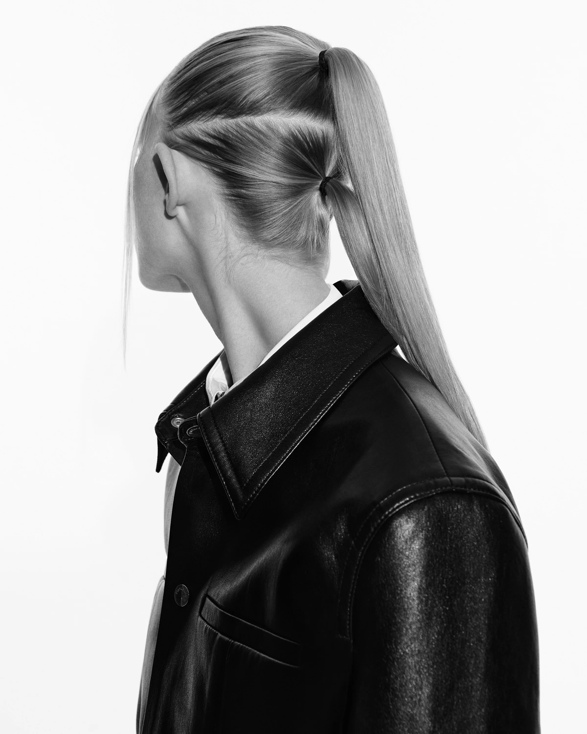 Black-and-white profile of a model with a sleek blonde split ponytail, wearing a black leather shirt jacket.
