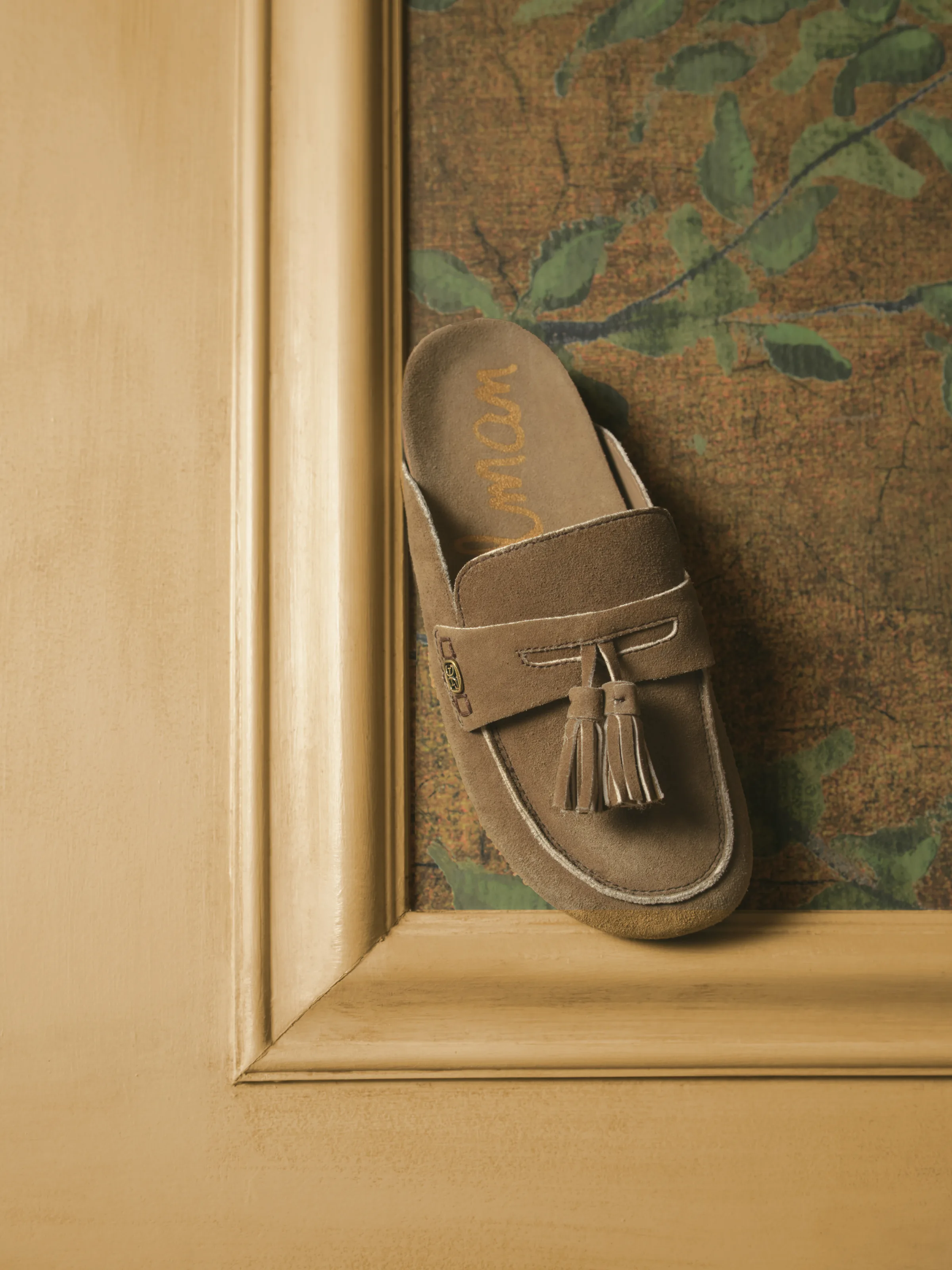 Tan suede tassel loafer mule perched on a gilded frame against a leafy tapestry backdrop, for Sam Edelman.