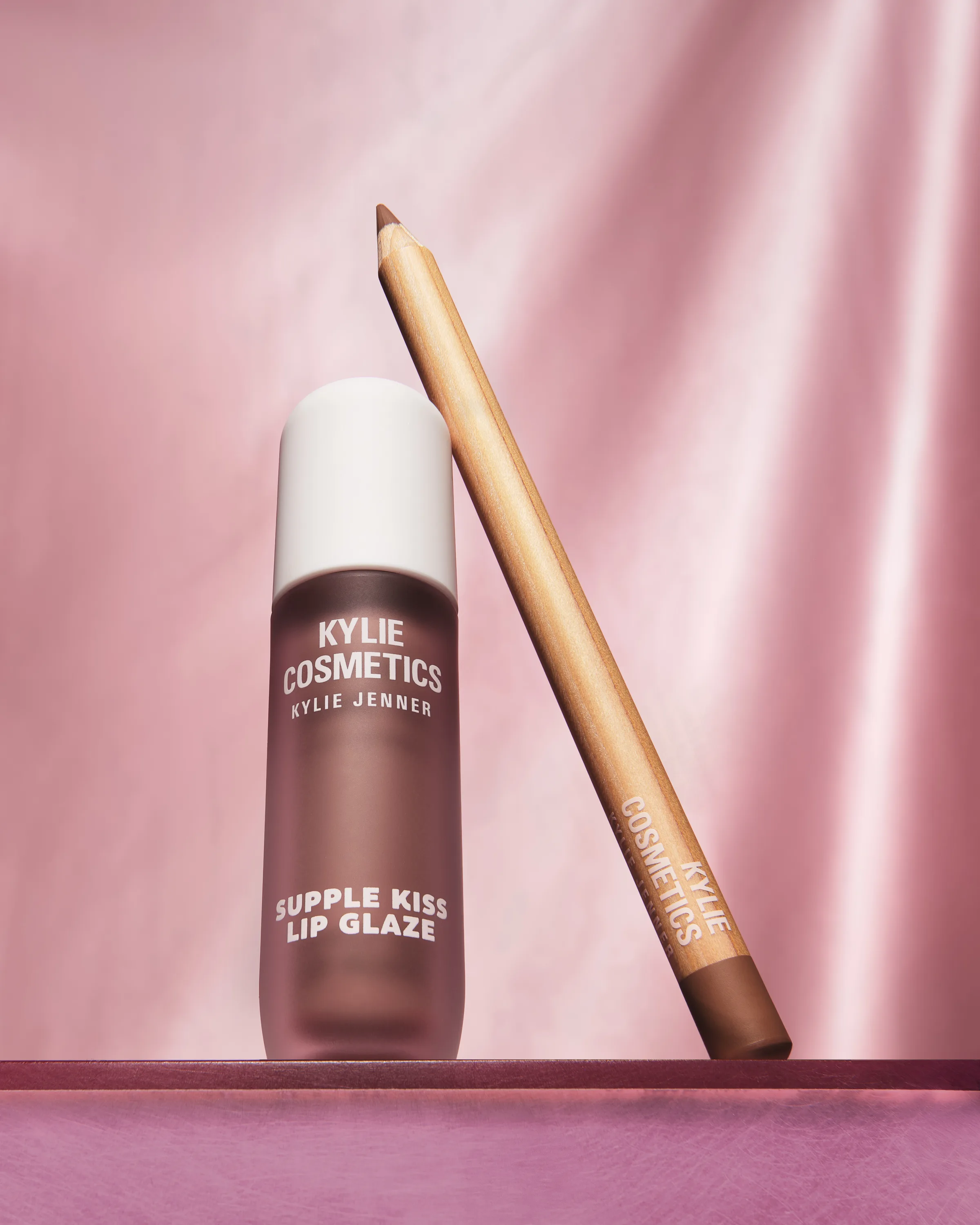 Supple Kiss Lip Glaze bottle and lip liner pencil on a pink ledge against draped pink satin, for Kylie Cosmetics.