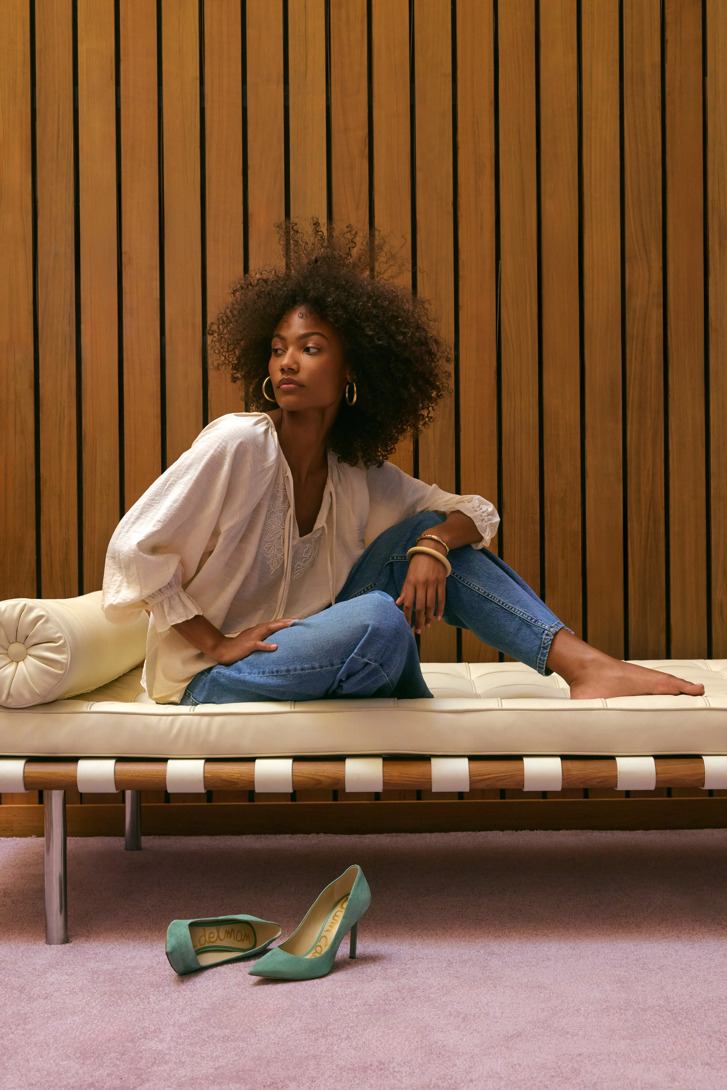 Model in white blouse and jeans on a cream leather daybed, green suede Hazel pumps on the floor, for Sam Edelman.