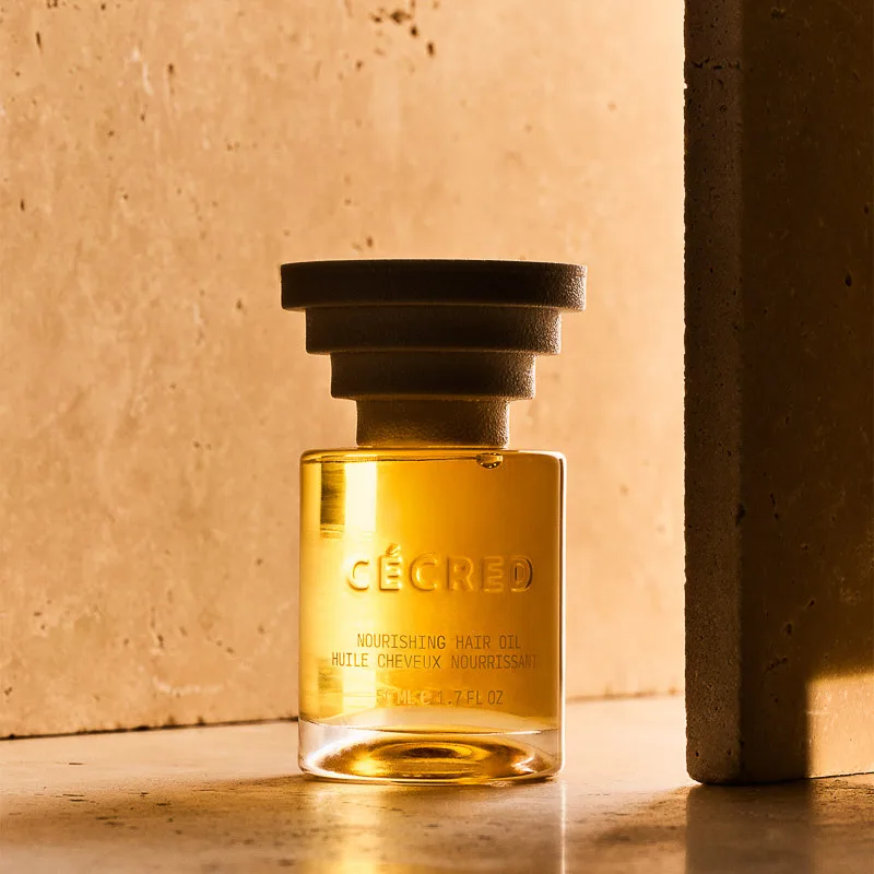 Glass bottle of nourishing hair oil with stacked cap on warm travertine stone in golden light, for Cecred.