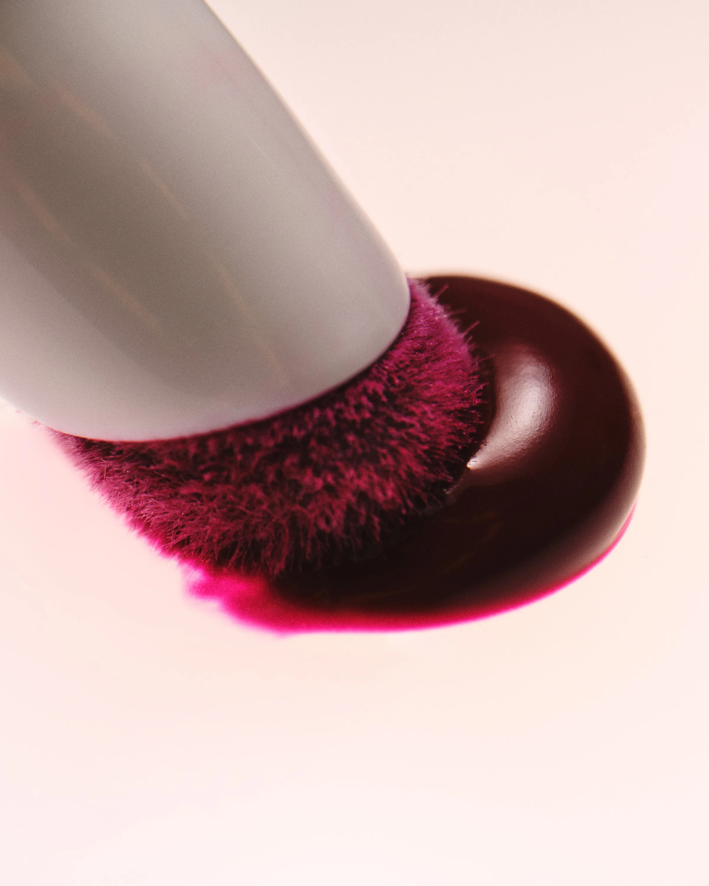Close-up of a white-handled makeup brush pressing into a glossy berry-red liquid pool, for Kylie Cosmetics.