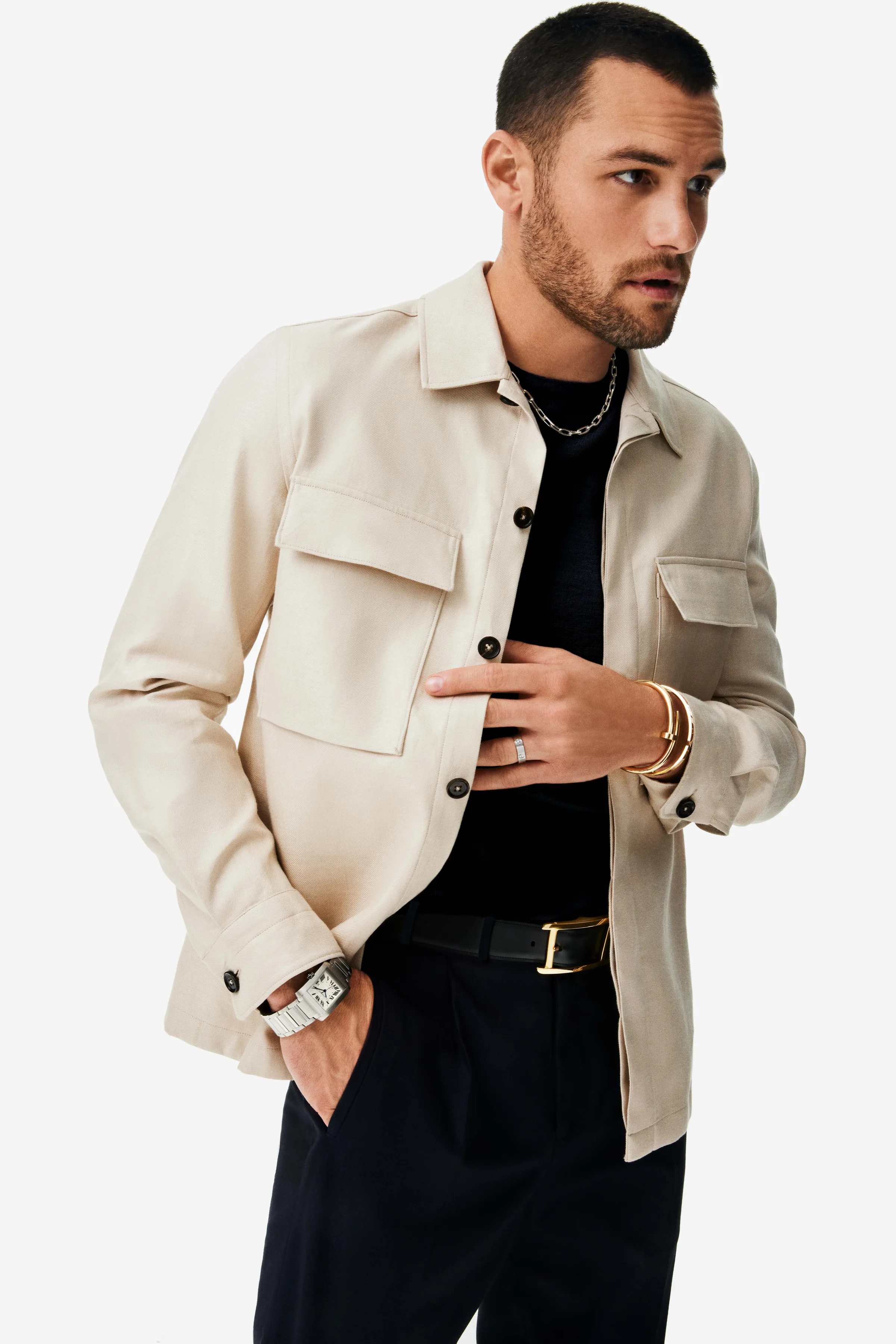 Man in a cream overshirt and black trousers wearing a Tank watch, gold bangles and silver chain on white, for Cartier.