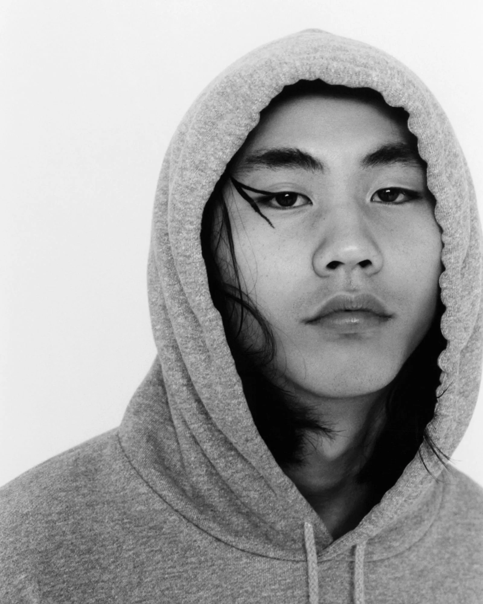 Black and white portrait of a model in a grey hoodie with long dark hair and graphic black eyeliner, for Vogue Korea.