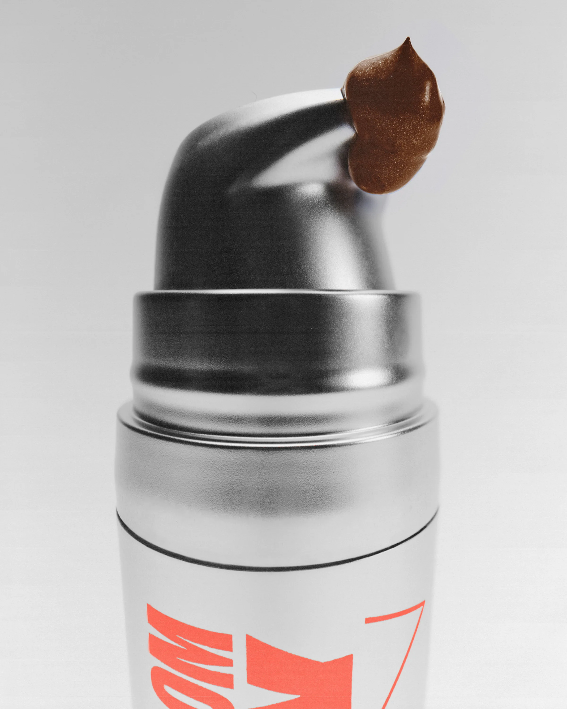 Close-up of a silver pump bottle with a dollop of bronze cream on the nozzle, coral lettering on the tube, for Good Weird.