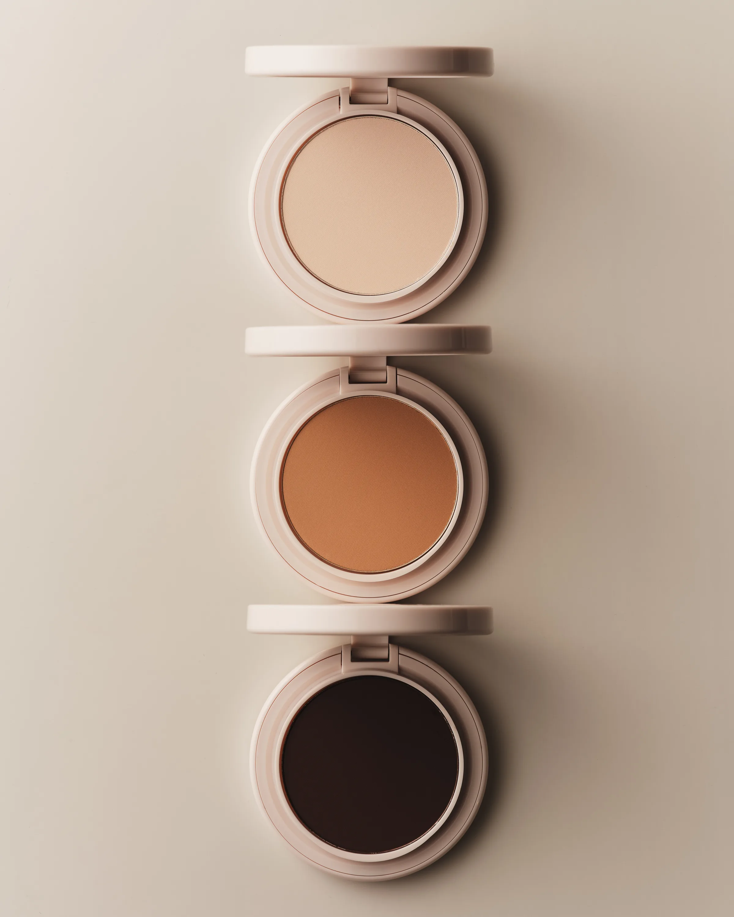 Three open pressed powder compacts in light, tan and deep shades stacked vertically on a beige backdrop, for Kylie Cosmetics.