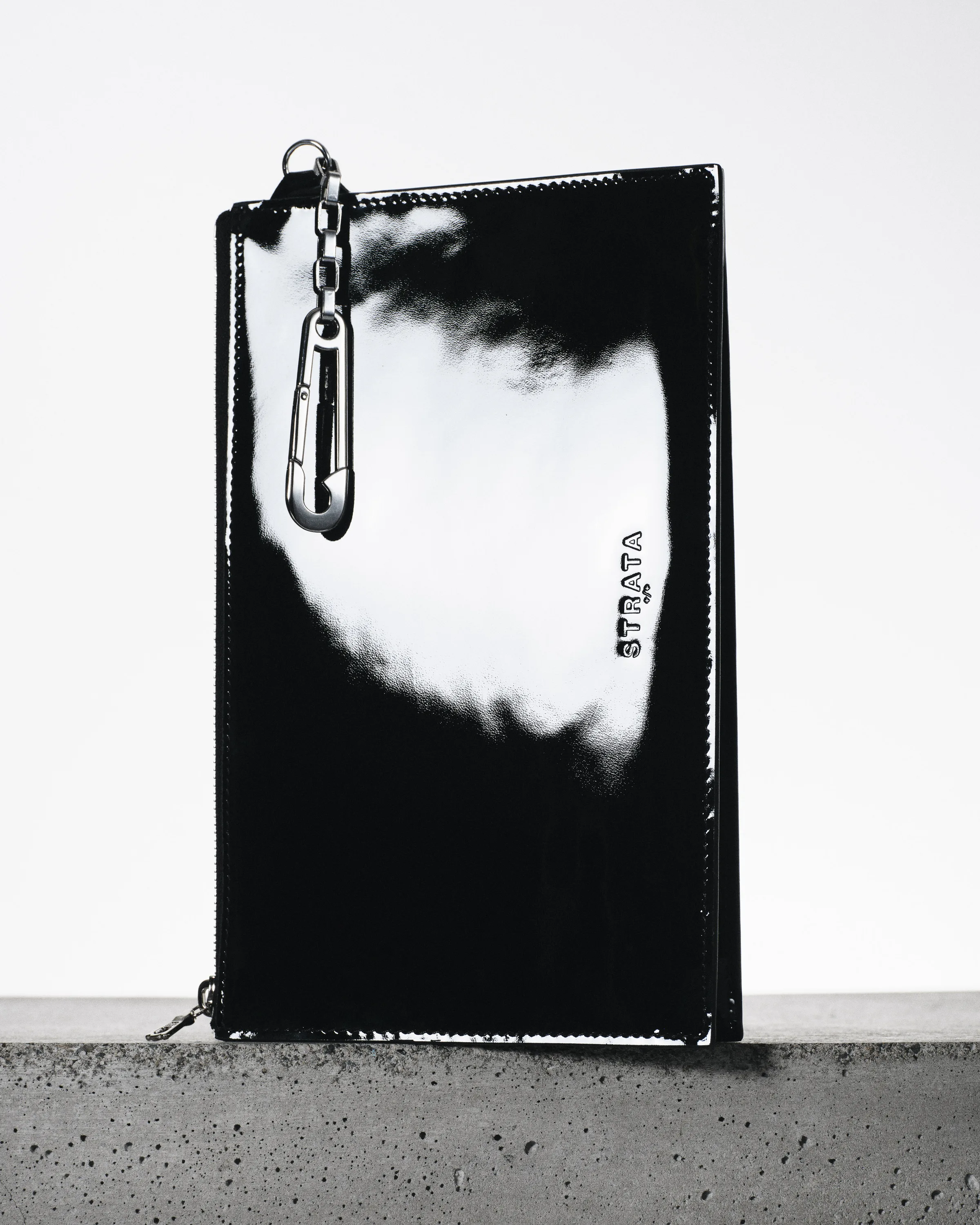 Black patent leather zip clutch with silver carabiner chain standing on a concrete ledge, for Strata.