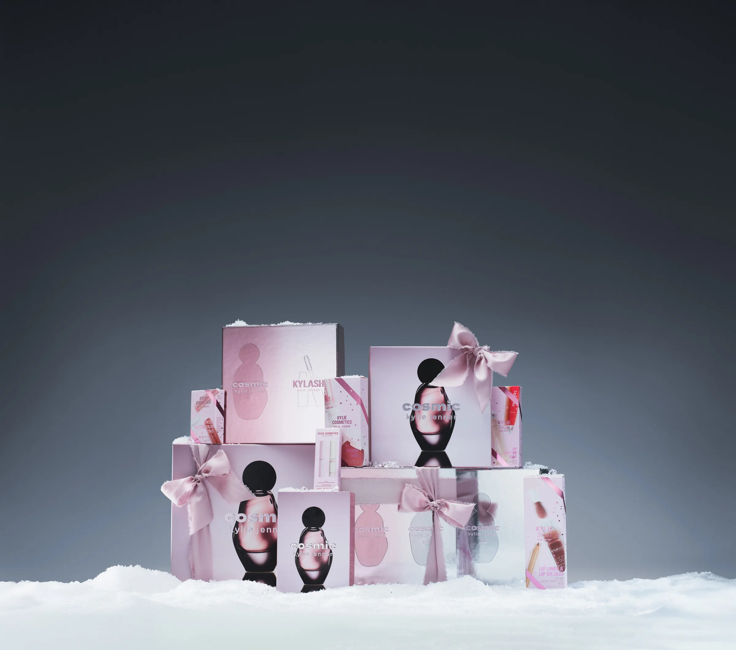 Stacked pink holiday gift sets with satin bows, Cosmic perfume and Kylash boxes on snow against grey, for Kylie Cosmetics.