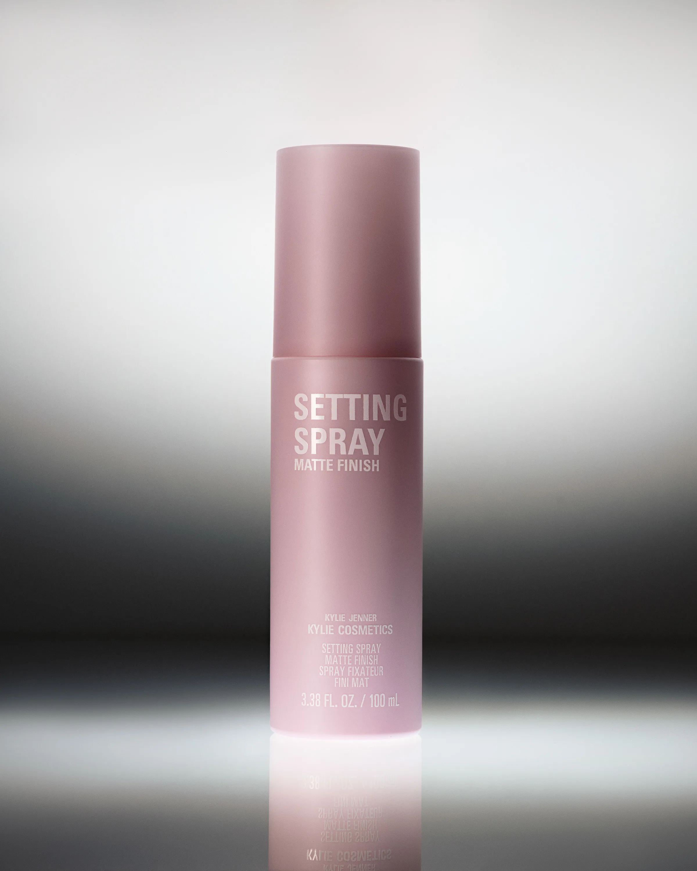 Pink matte-finish setting spray bottle on a reflective surface against a silver gradient backdrop, for Kylie Cosmetics.