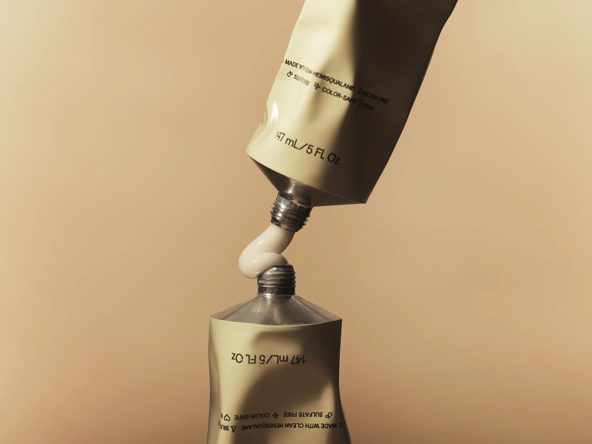 Two crumpled sage-green tubes, one inverted, squeezing white cream into each other on a warm beige backdrop, for JVN.