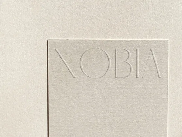 Cream textured paper card with the NOBIA wordmark blind-debossed across the top, on matching paper, for Nobia.