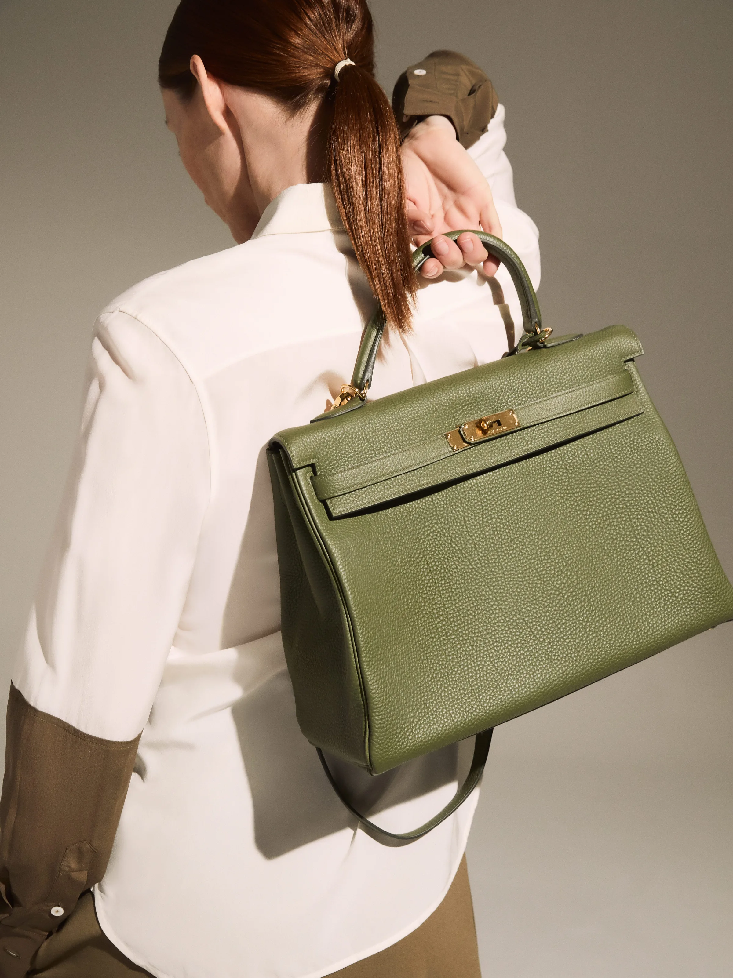 Model in a white shirt holds a green pebbled leather handbag with gold hardware over her shoulder, for The RealReal.