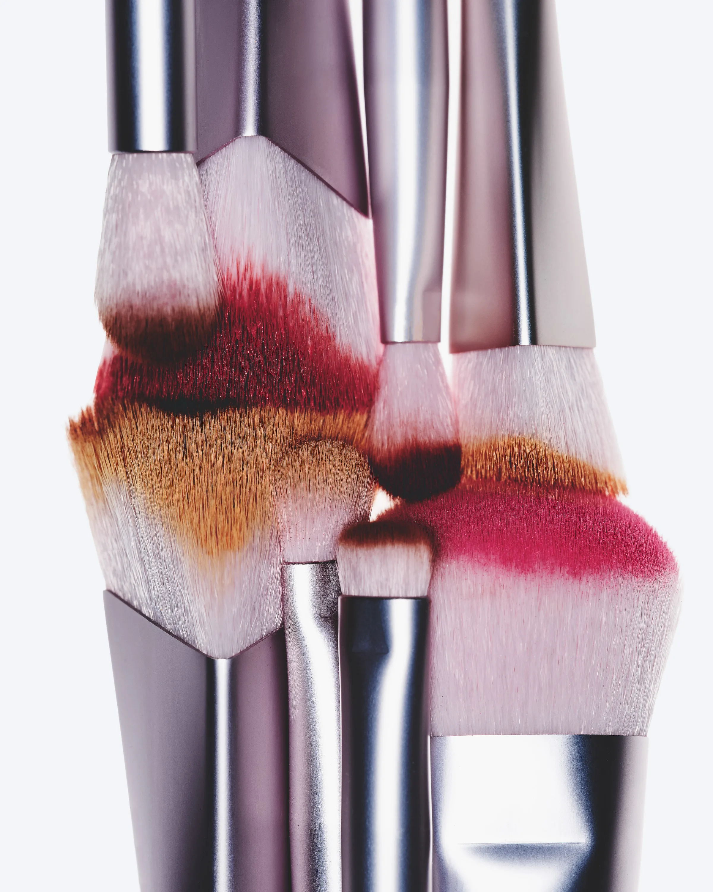 Makeup brushes with chrome ferrules pressed together, bristles stained red, pink and bronze pigment, for Kylie Cosmetics.
