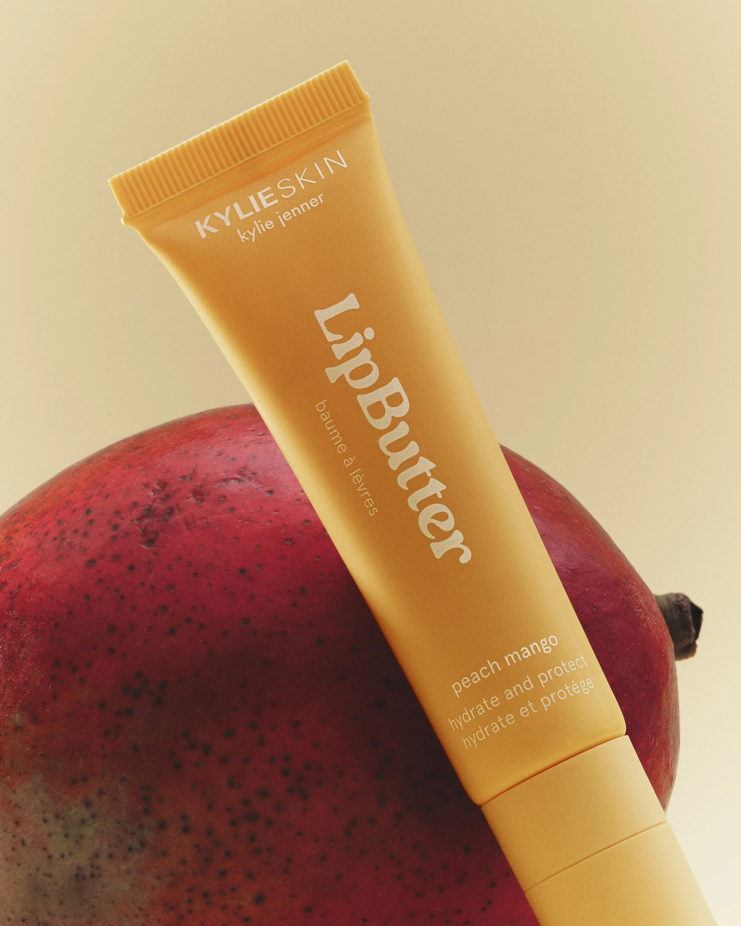 Peach mango Lip Butter tube leaning on a red mango against a warm beige backdrop, for Kylie Skin.