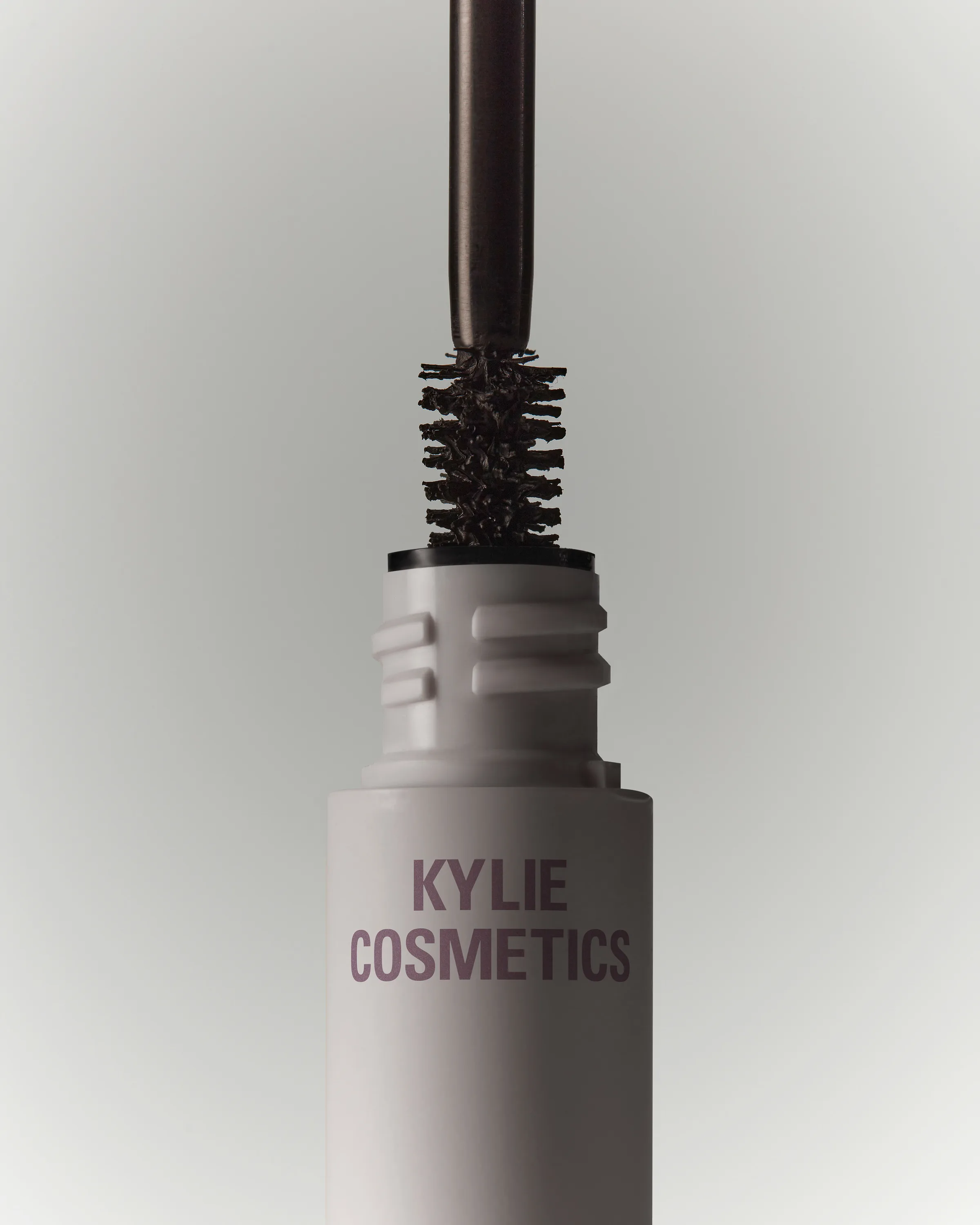 Mascara wand lifted from a pale grey tube, dark bristles coated in formula, on a grey backdrop, for Kylie Cosmetics.