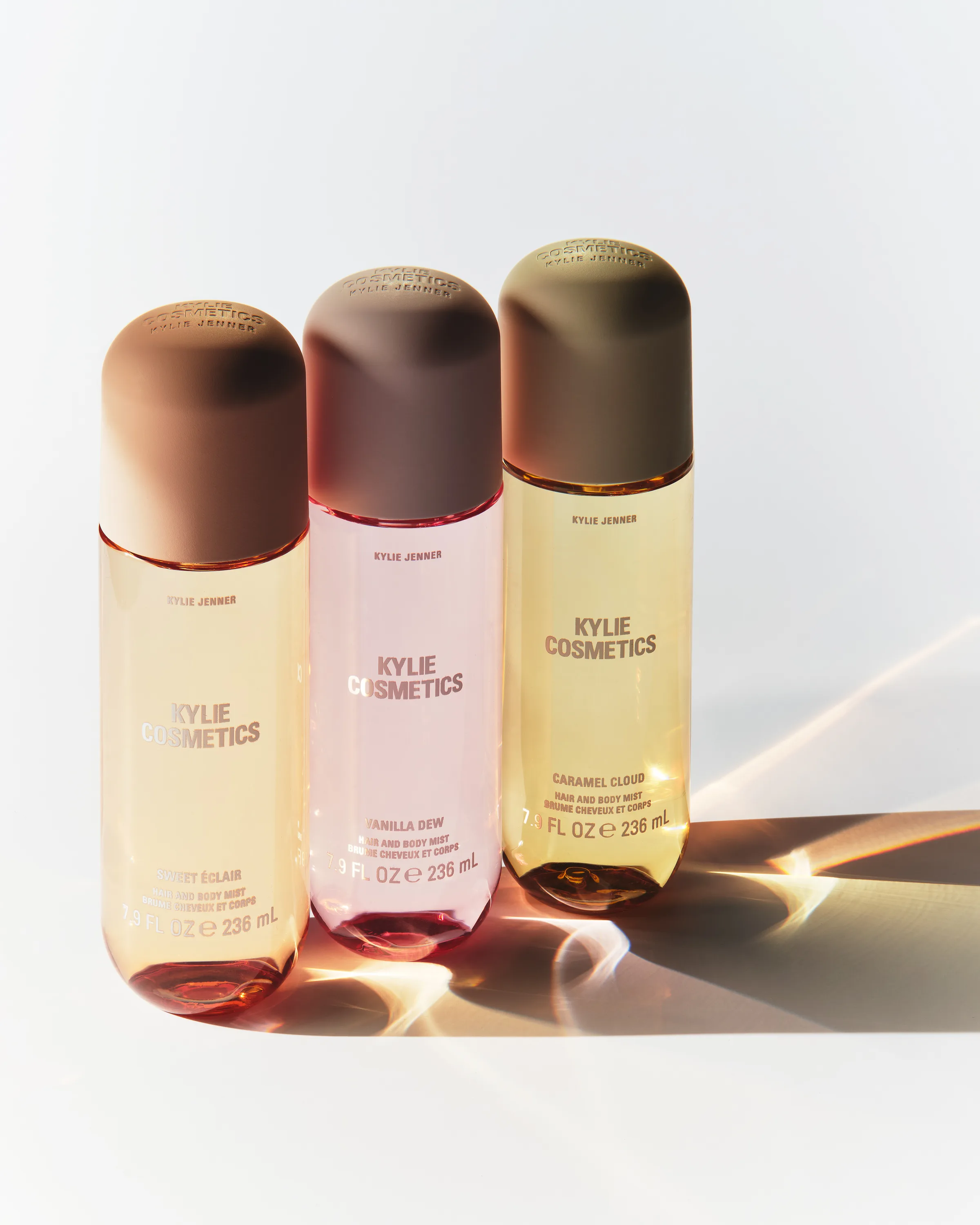 Three hair and body mist bottles in Sweet Eclair, Vanilla Dew and Caramel Cloud casting sunlit shadows, for Kylie Cosmetics.