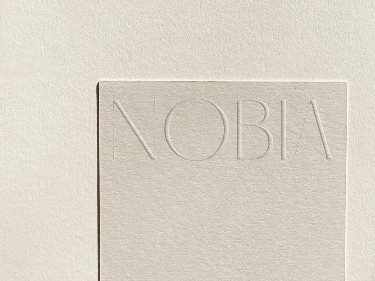 Cream textured paper card with the NOBIA wordmark blind-debossed across the top, on matching paper, for Nobia.