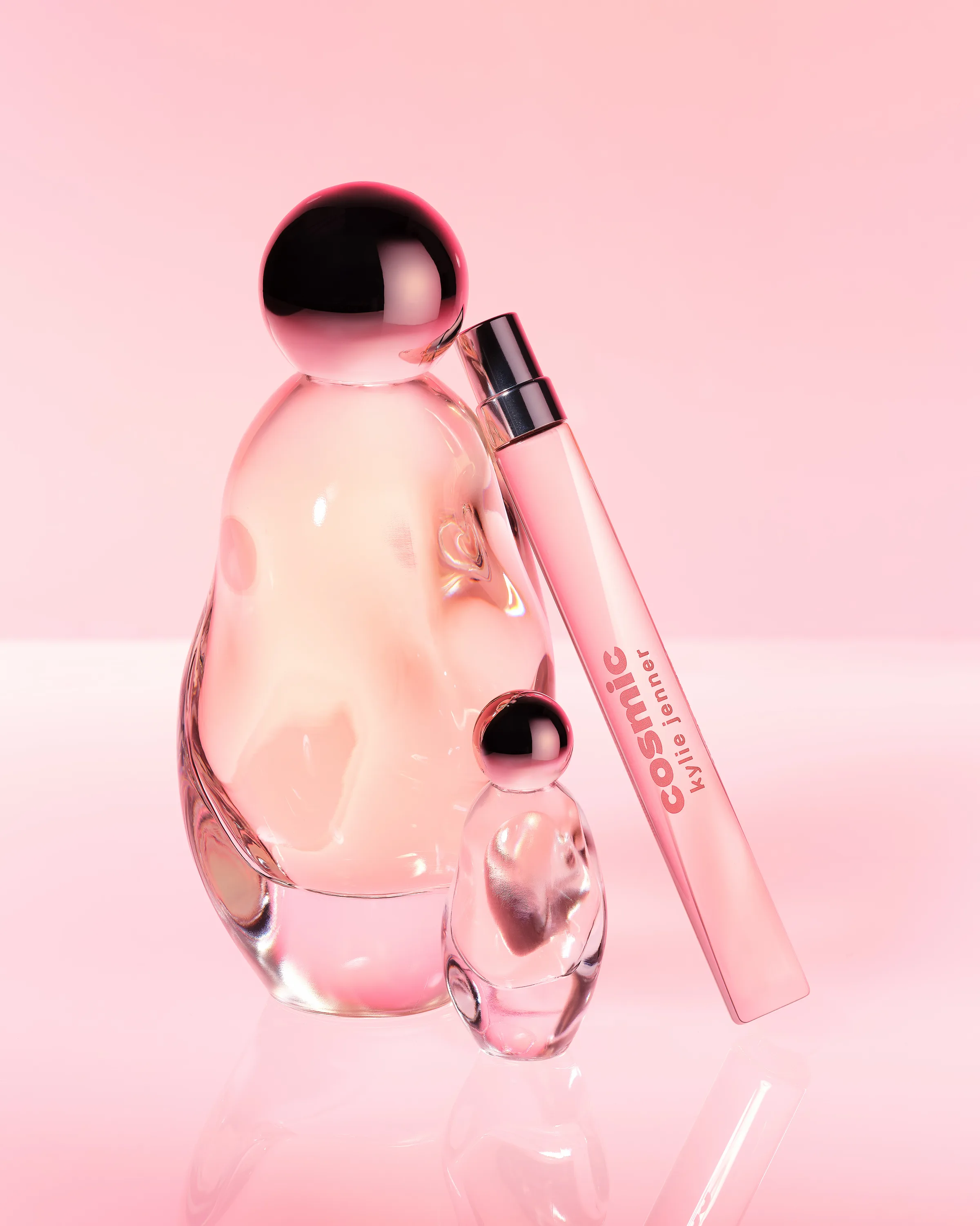 Still life of Cosmic perfume bottle, travel spray and mini on a glossy pink surface, for Kylie Cosmetics.
