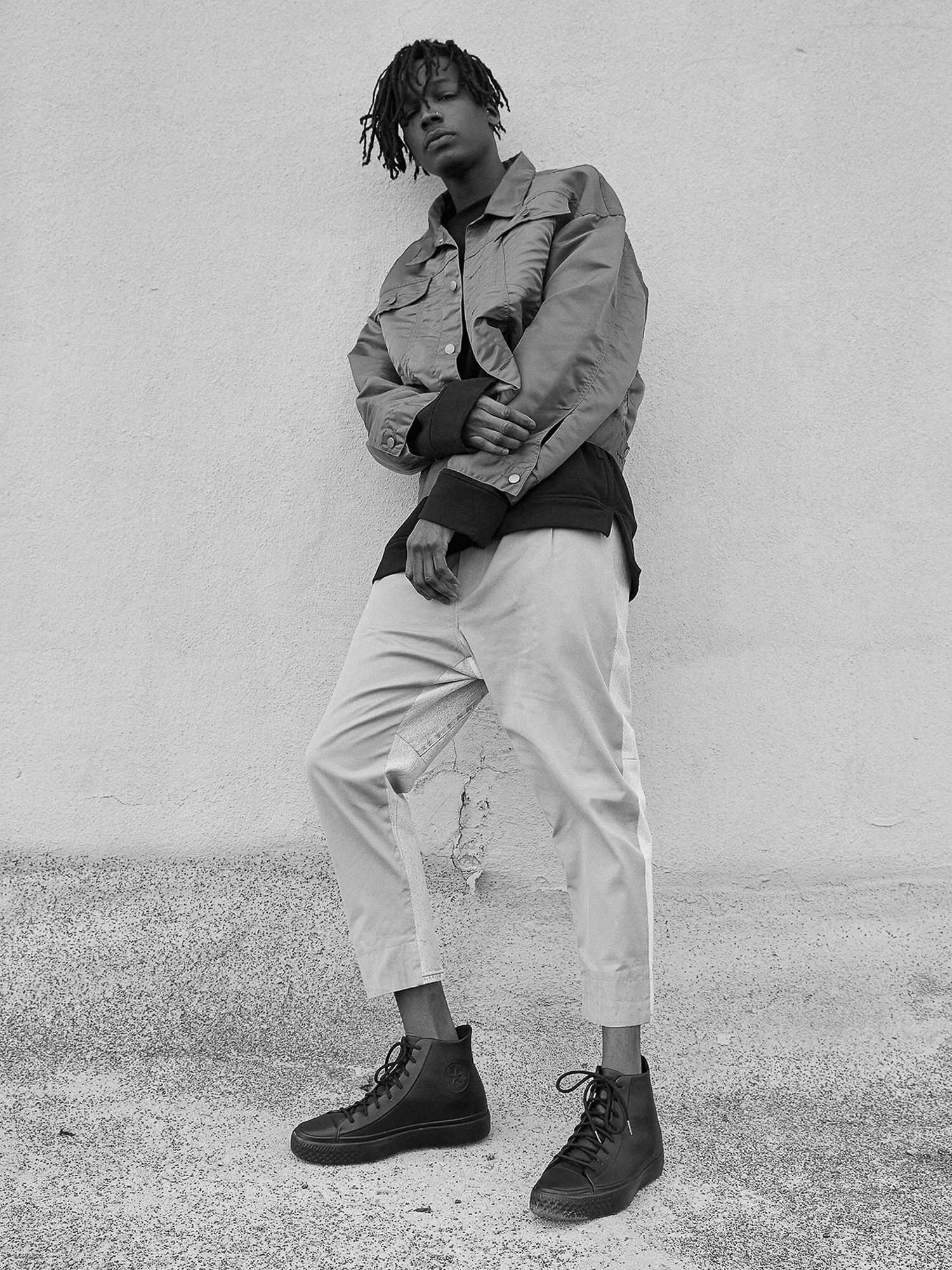 Black and white shot of a model leaning on a wall in a satin jacket, cropped trousers and black high-top sneakers, for Converse.
