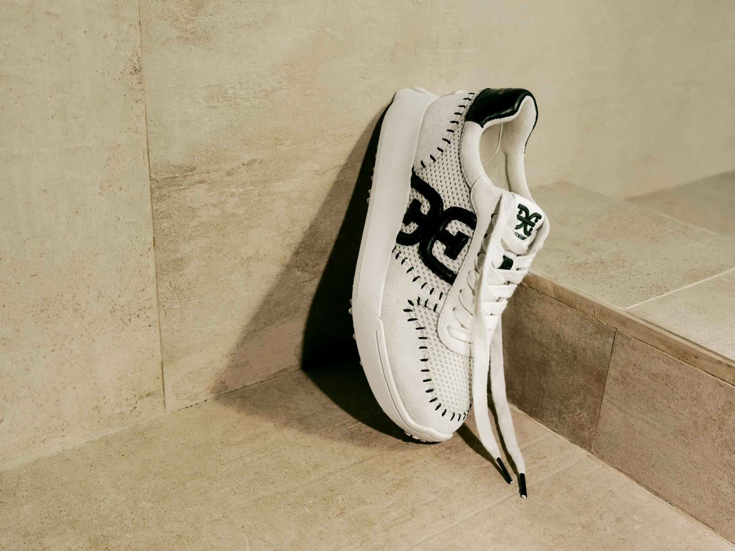 White knit sneaker with black double-E logo and whipstitch trim leaning against a beige stone wall, for Sam Edelman.