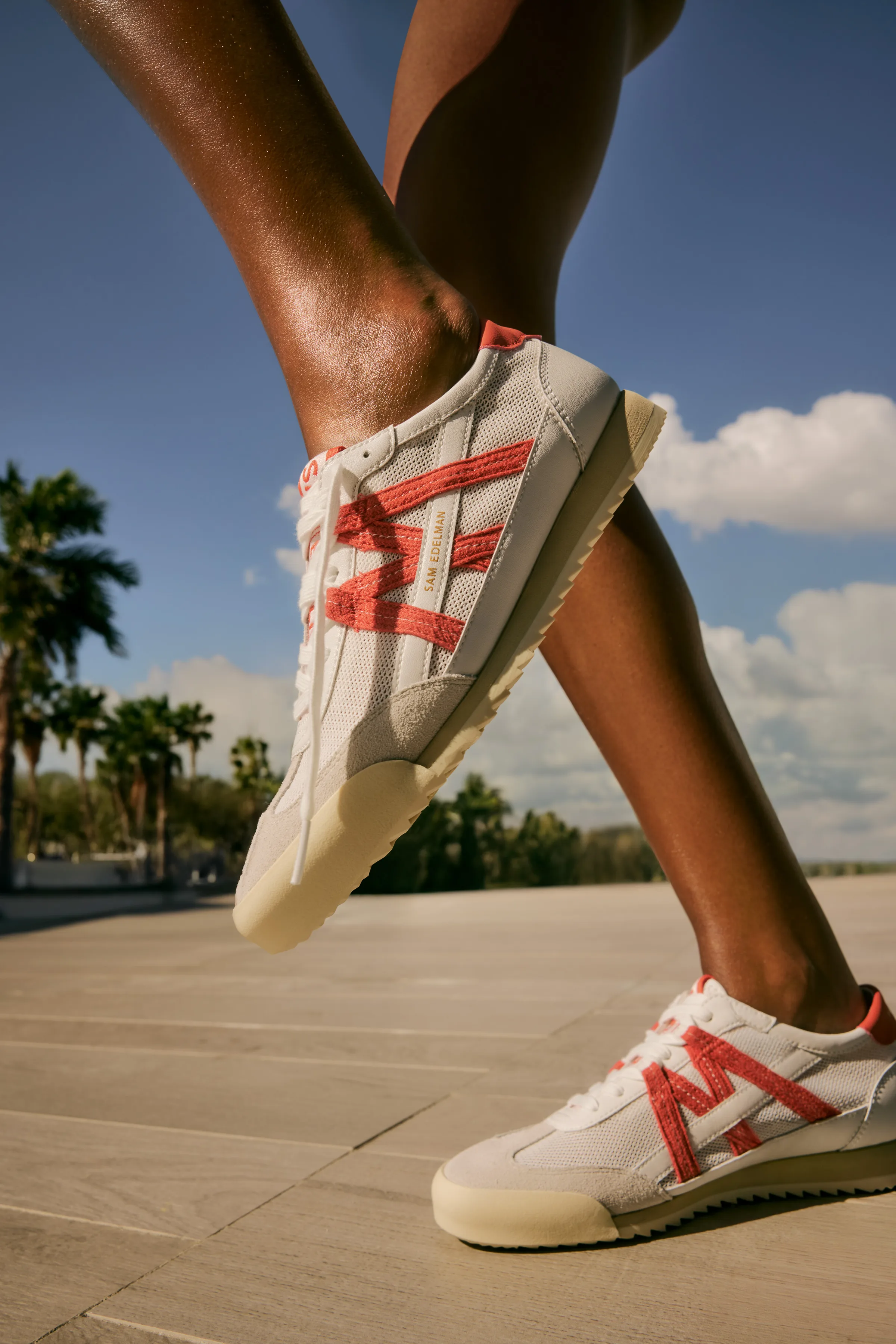 Close-up of legs mid-stride in white mesh sneakers with coral stripes on a sunny plaza with palm trees, for Sam Edelman.
