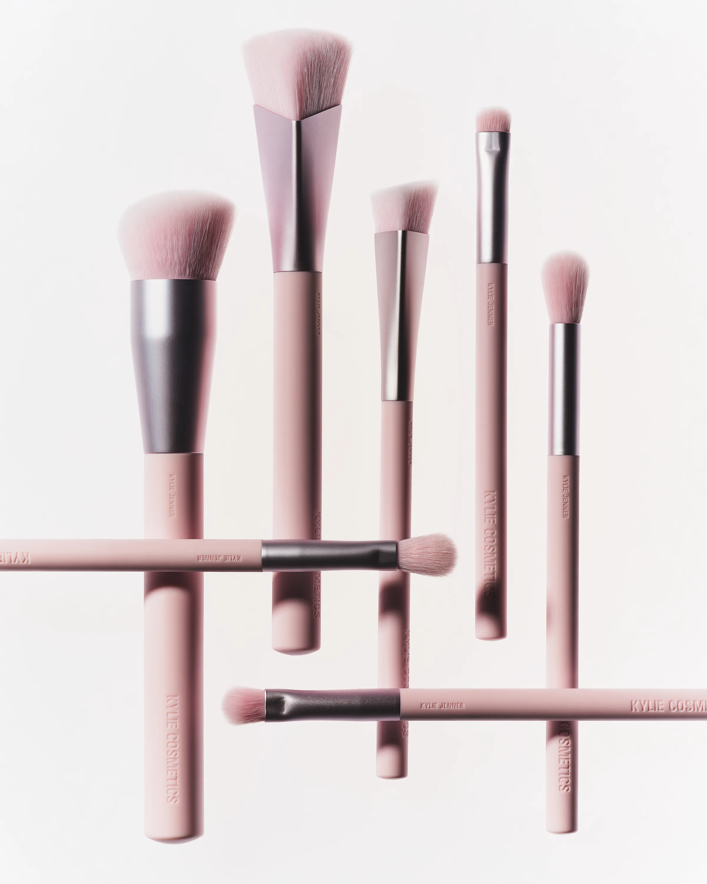 Seven pink makeup brushes with silver ferrules arranged vertically and horizontally on white, for Kylie Cosmetics.