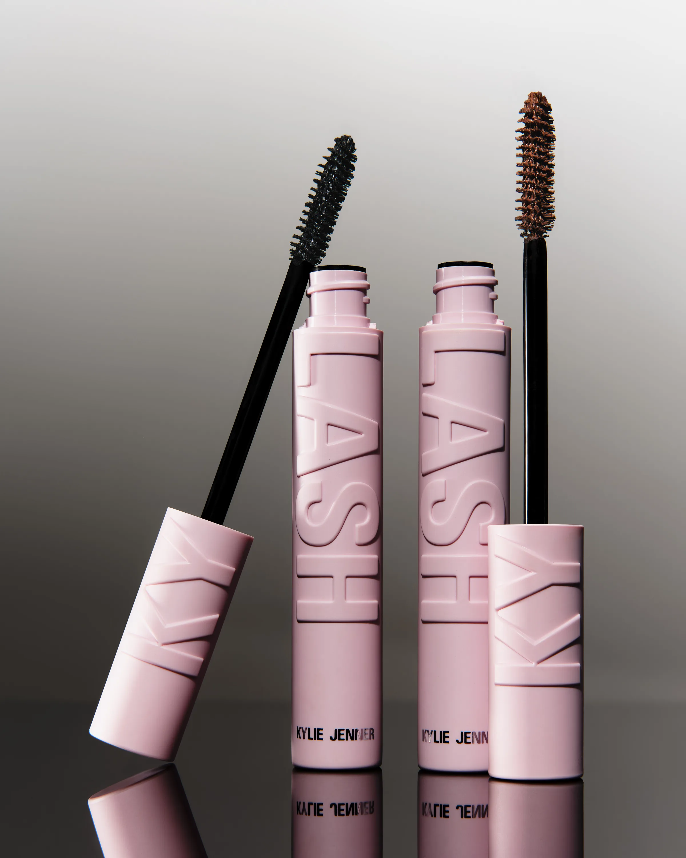 Two pink Kylash mascara tubes with black and brown wands on a reflective grey gradient, for Kylie Cosmetics.