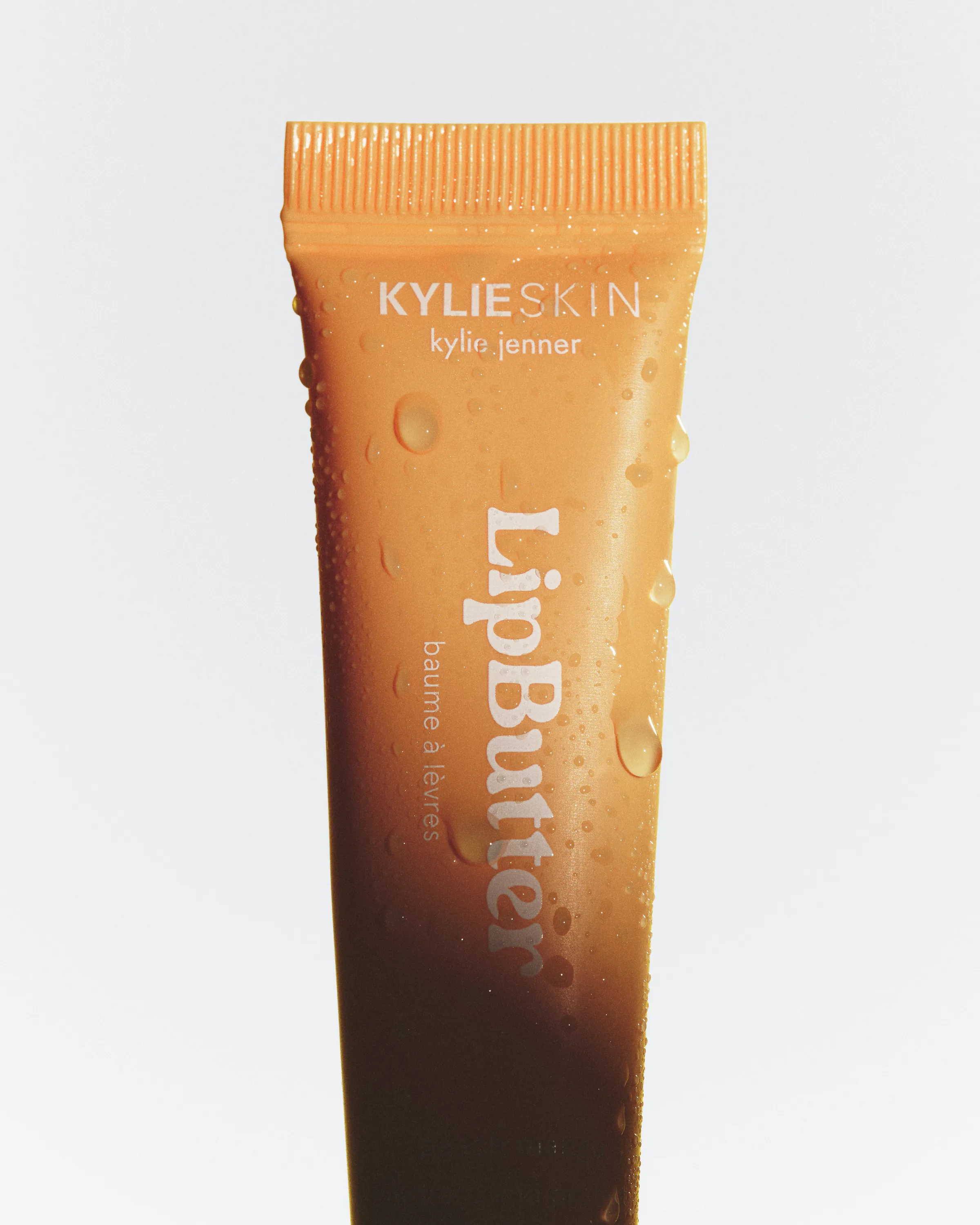 Orange Lip Butter tube covered in water droplets against a pale grey background, for Kylie Skin.