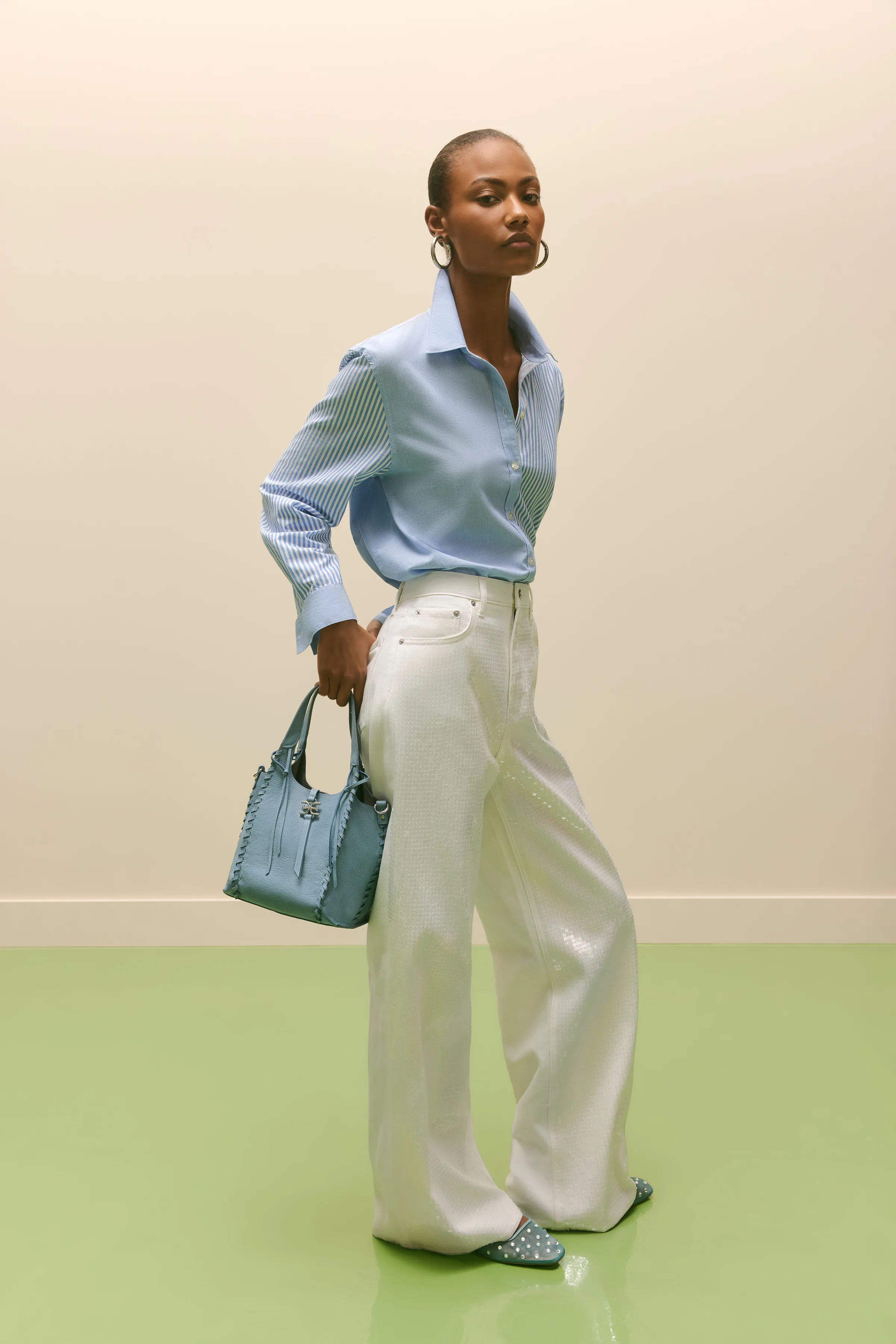 Model in striped blue shirt and white sequin trousers holding a blue leather handbag on a green floor, for Sam Edelman.