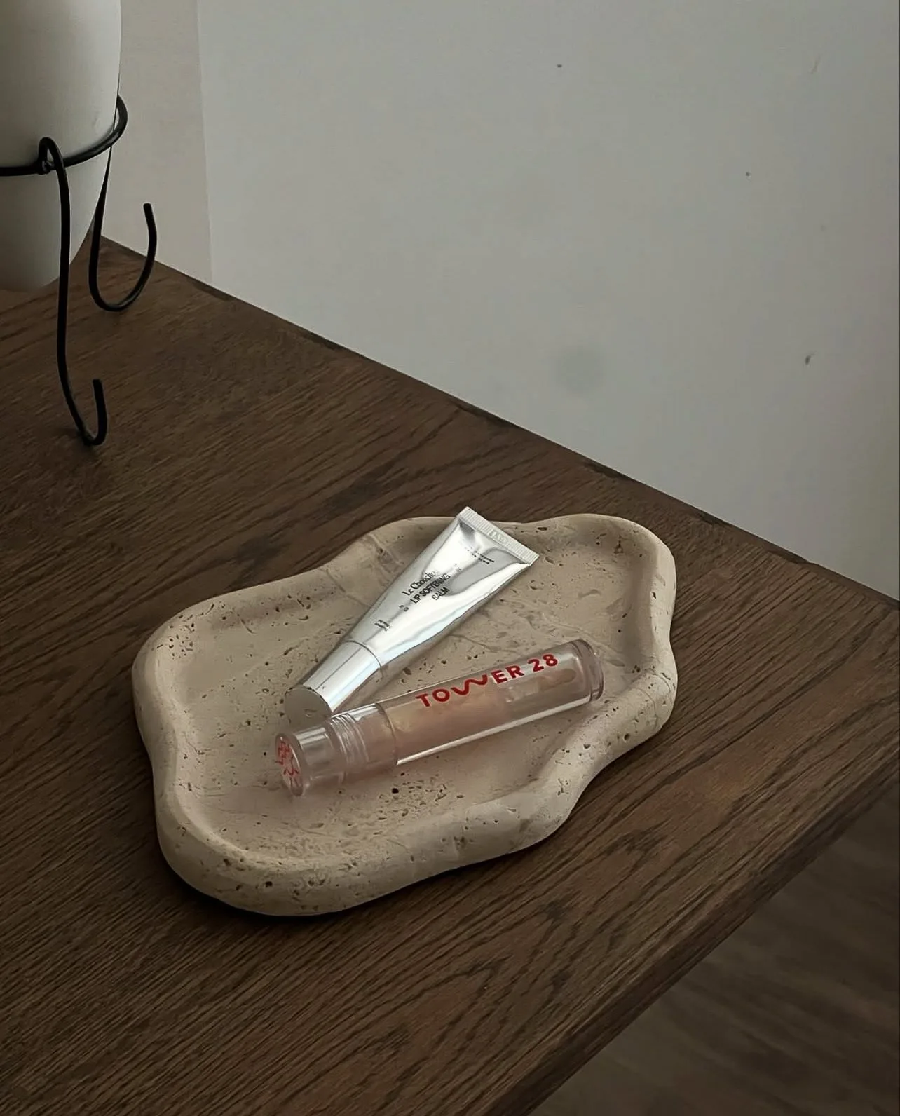 Tower 28 lip gloss and a silver lip balm tube on a wavy travertine tray atop a wooden table, for Tower 28.