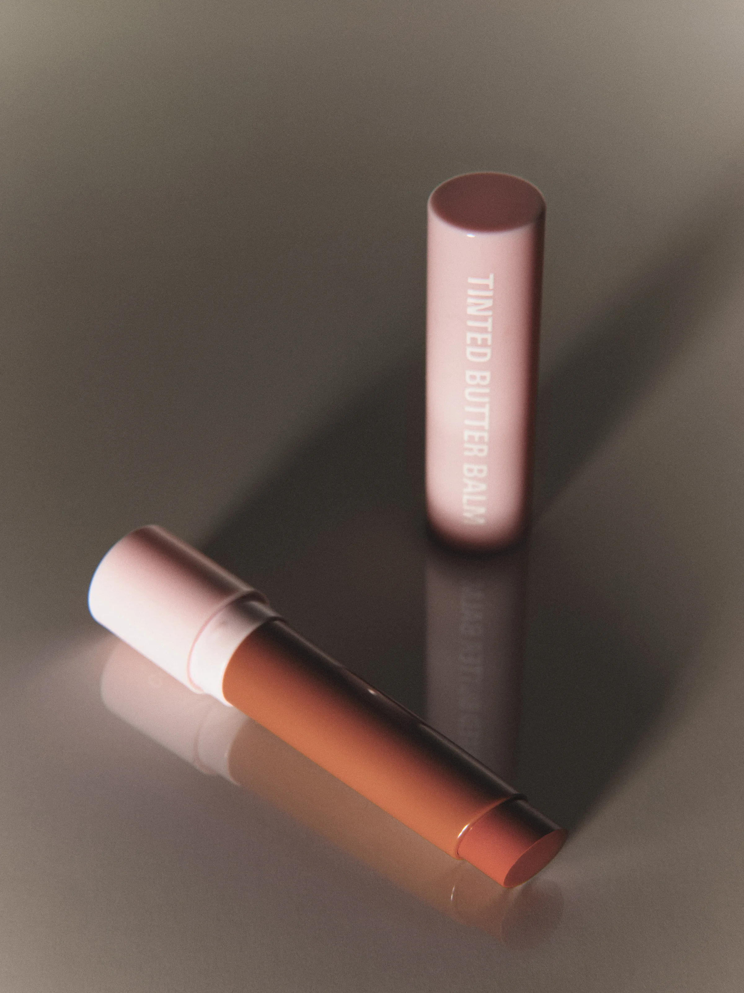 Pink Tinted Butter Balm tube standing beside an open honey-toned balm on a glossy taupe surface, for Kylie Cosmetics.