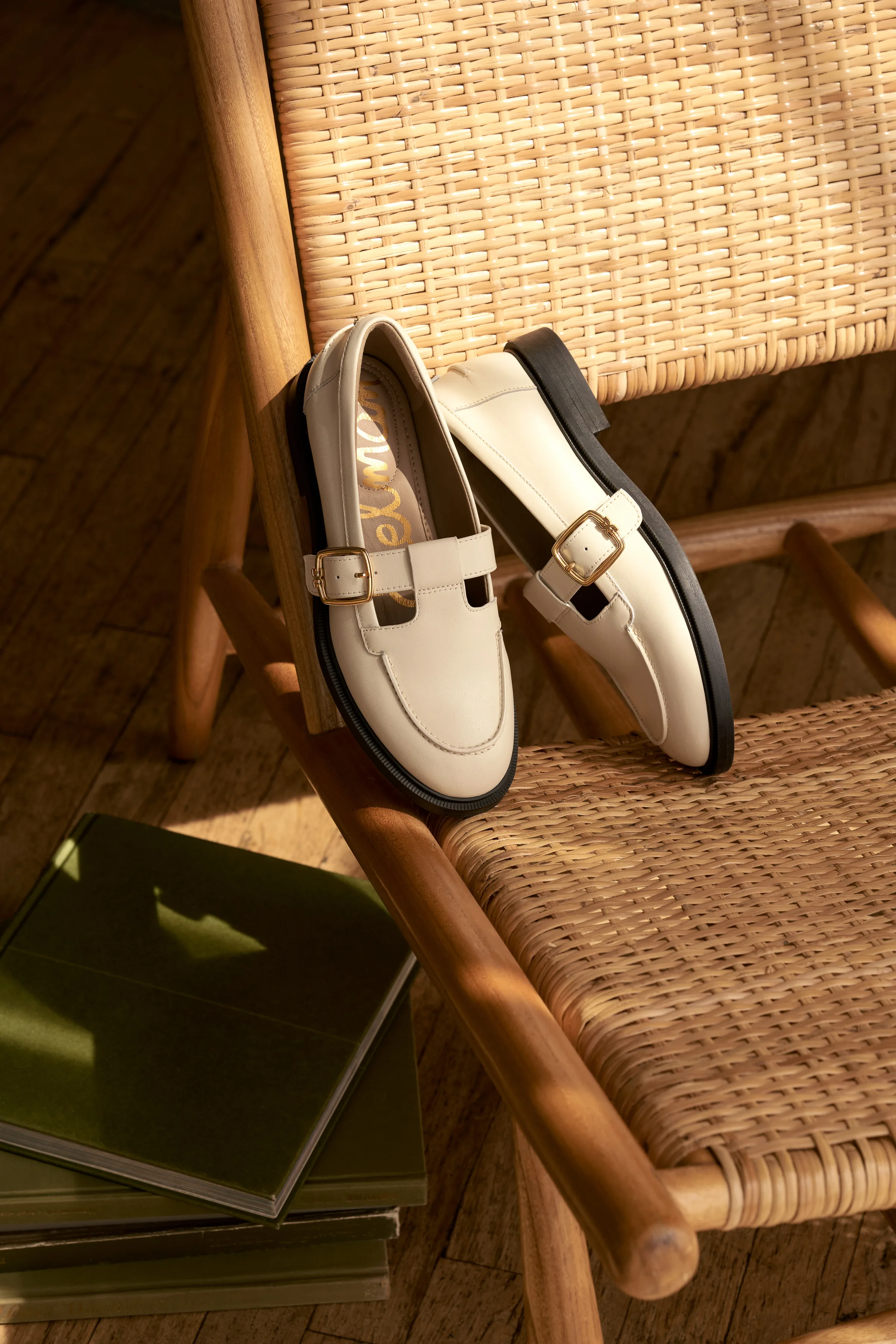 Cream leather loafers with gold buckle straps resting on a wooden cane chair in warm sunlight, for Sam Edelman.