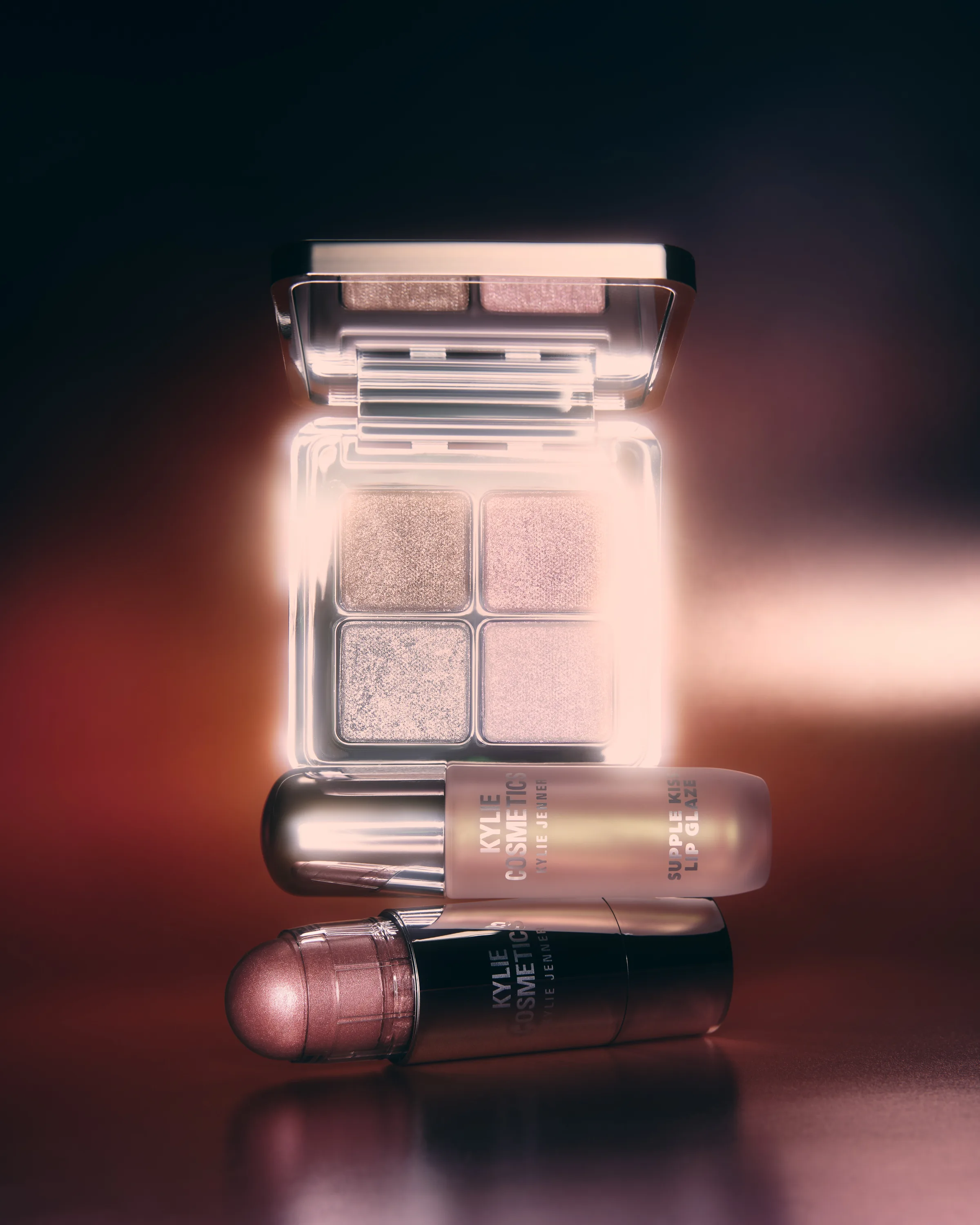 Still life of a glowing eyeshadow palette stacked on a lip glaze and rose balm stick against a warm red backdrop, for Kylie Cosmetics.