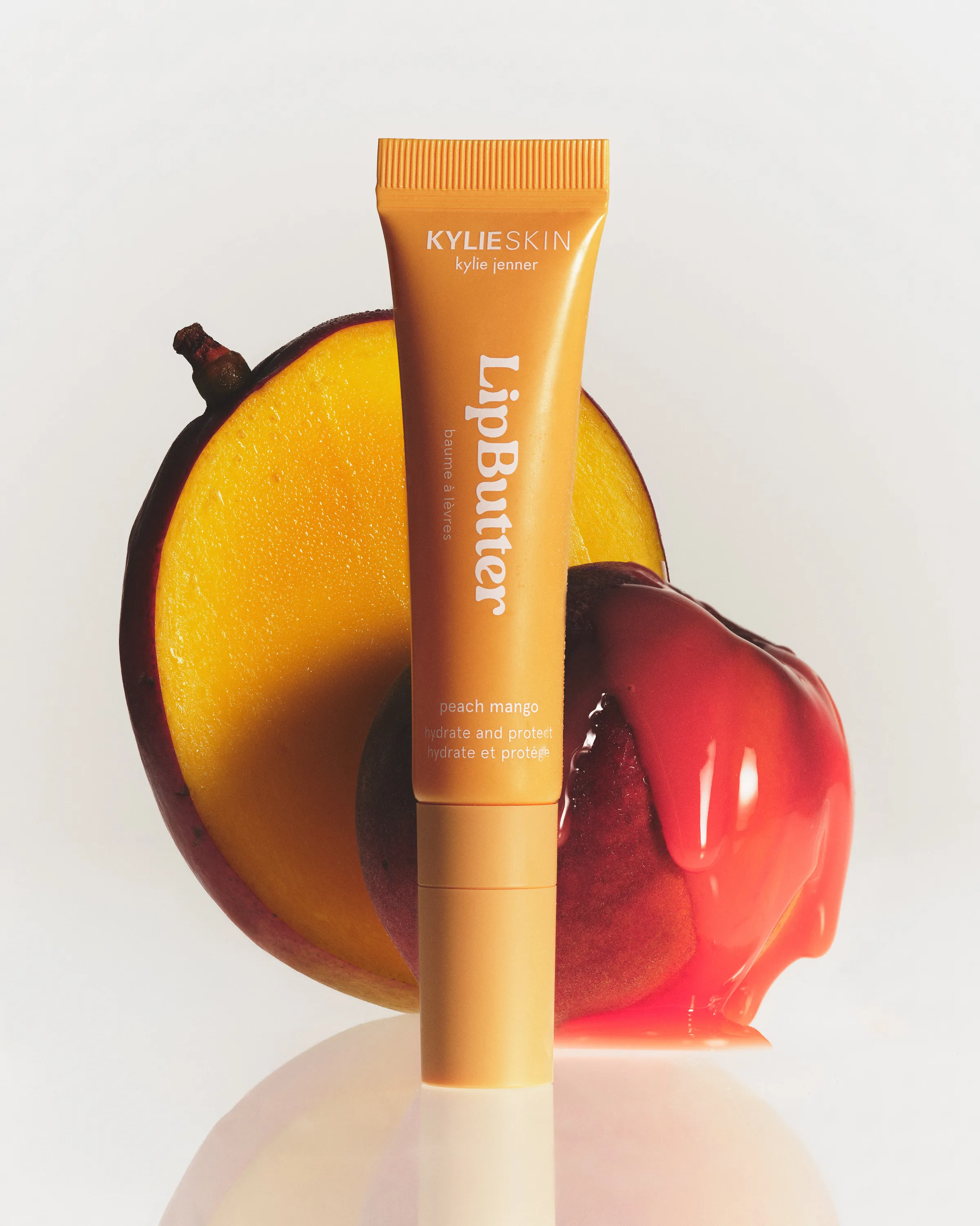 Peach mango Lip Butter tube in front of a halved mango and a peach dripping glossy red balm, for Kylie Skin.