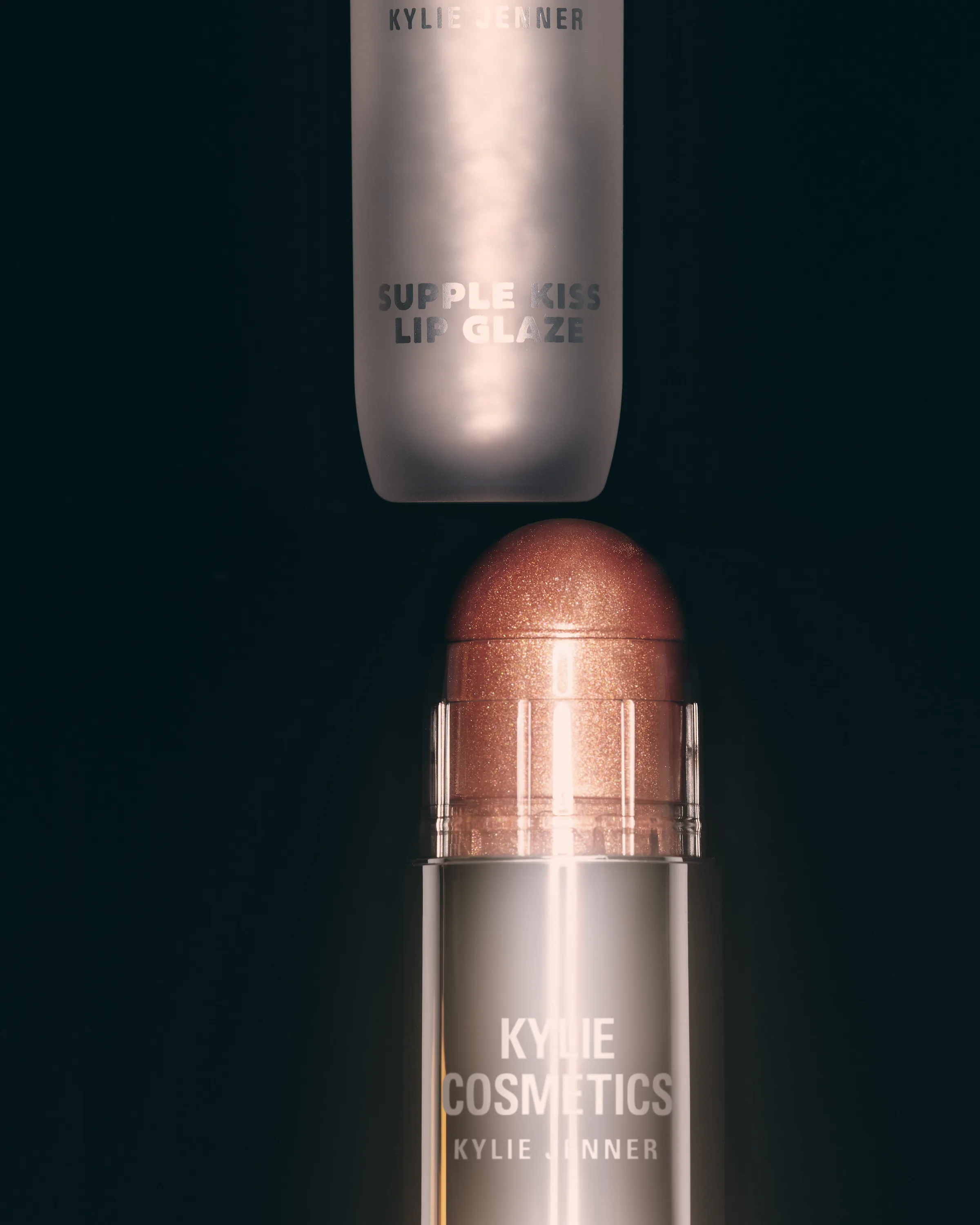 Supple Kiss Lip Glaze tube above a shimmering peach balm stick on a black background, for Kylie Cosmetics.
