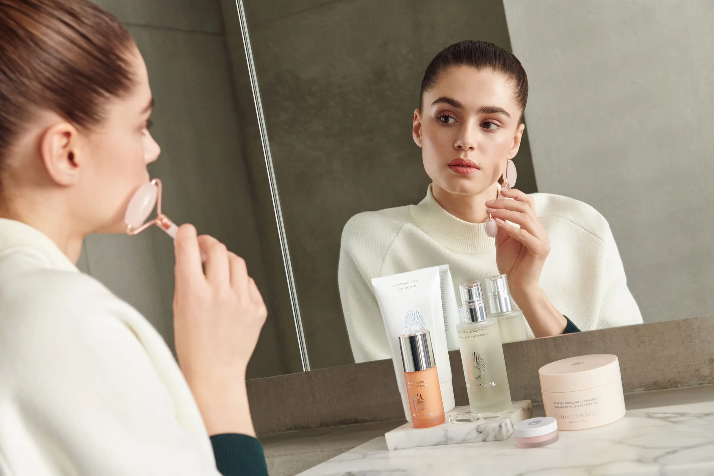 Woman using a rose quartz facial roller at a mirror beside skincare bottles on a marble vanity, for Net-a-Porter.