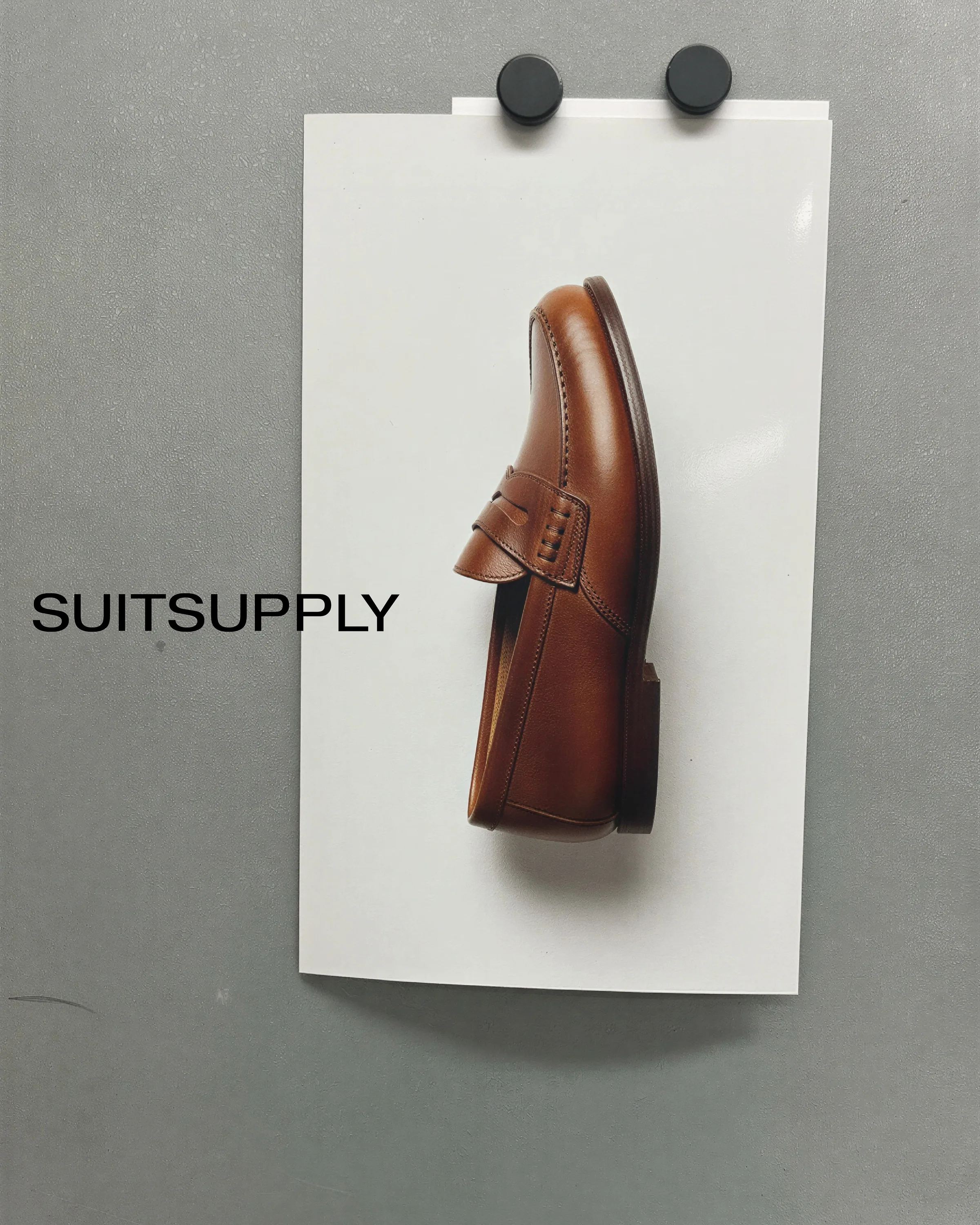 AI image of a tan leather penny loafer on a white print held by magnets to a grey wall. Suitsupply AI Concept.
