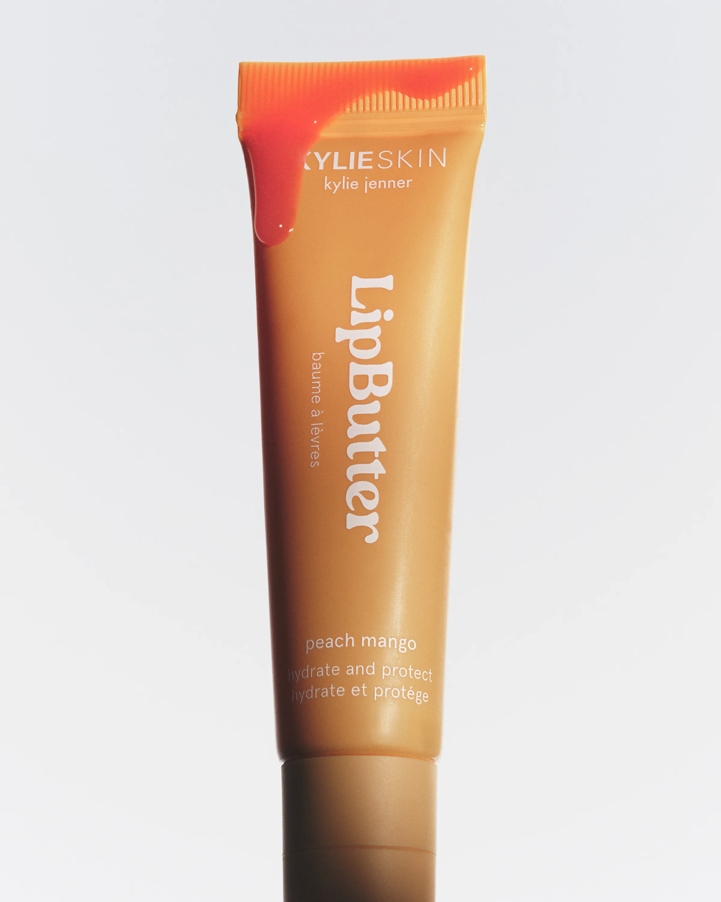 Peach mango Kylie Skin Lip Butter tube with glossy balm dripping over its cap on a white background, for Kylie Cosmetics.