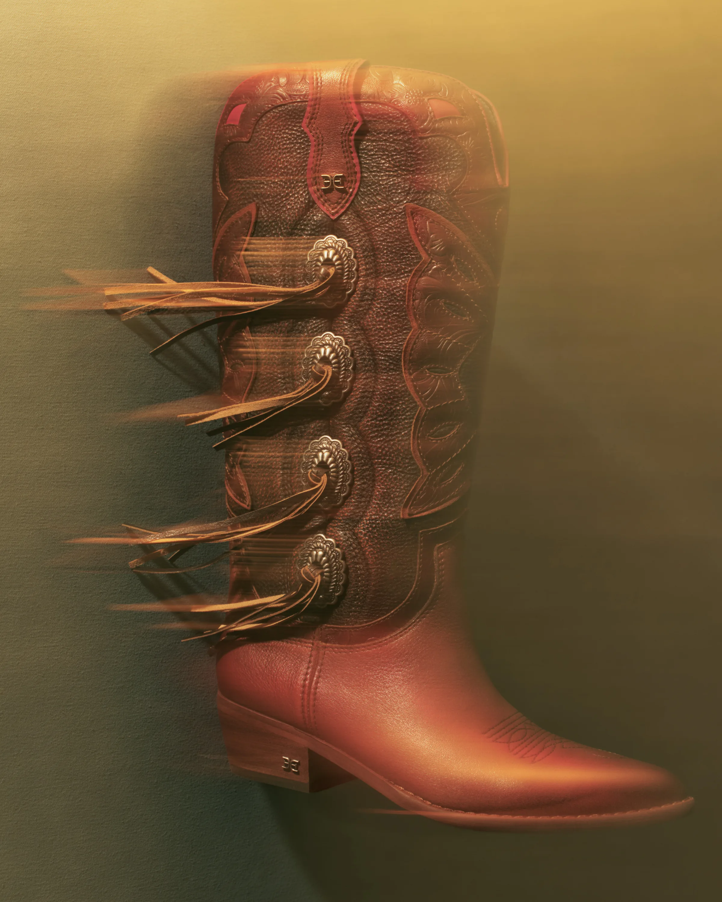 Brown leather western boot with concho buttons and motion-blurred fringe on a warm gradient backdrop, for Sam Edelman.